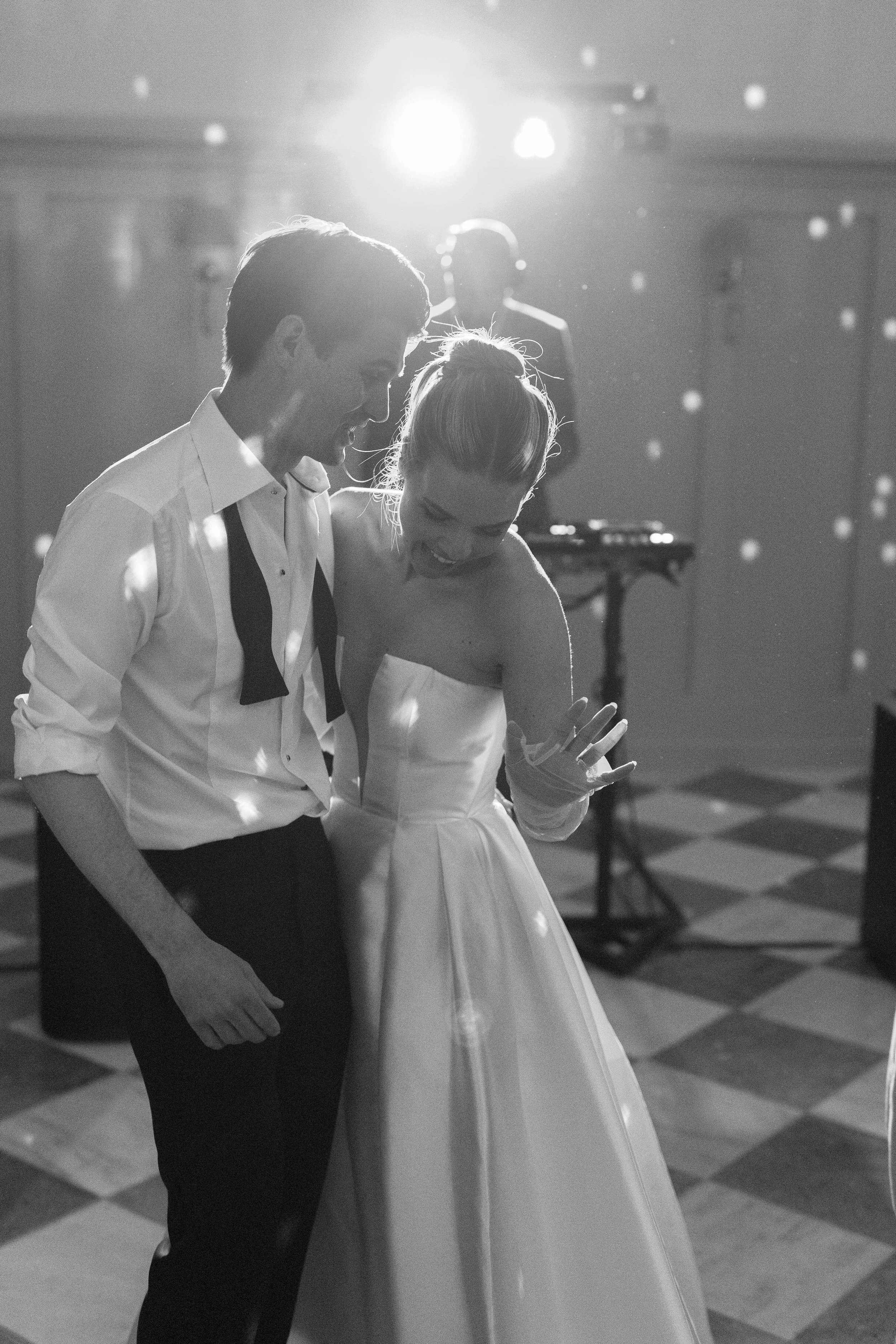 Black and white photo of a bride and groom dancing at their wedding reception, smiling and enjoying the moment with a DJ in the background.