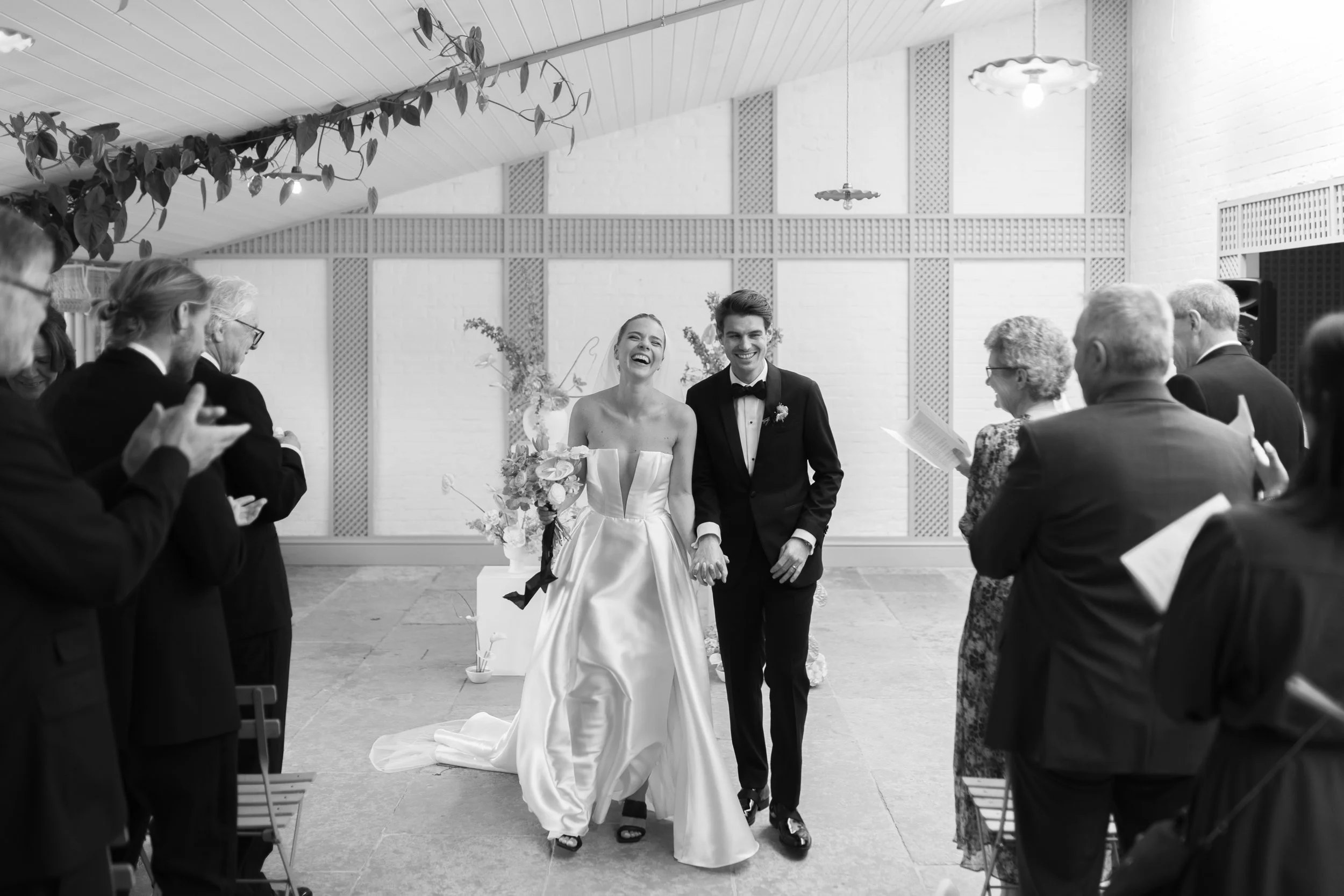 A bride and groom walking down the aisle, smiling and holding hands, surrounded by seated guests during a wedding ceremony in a decorated indoor venue.