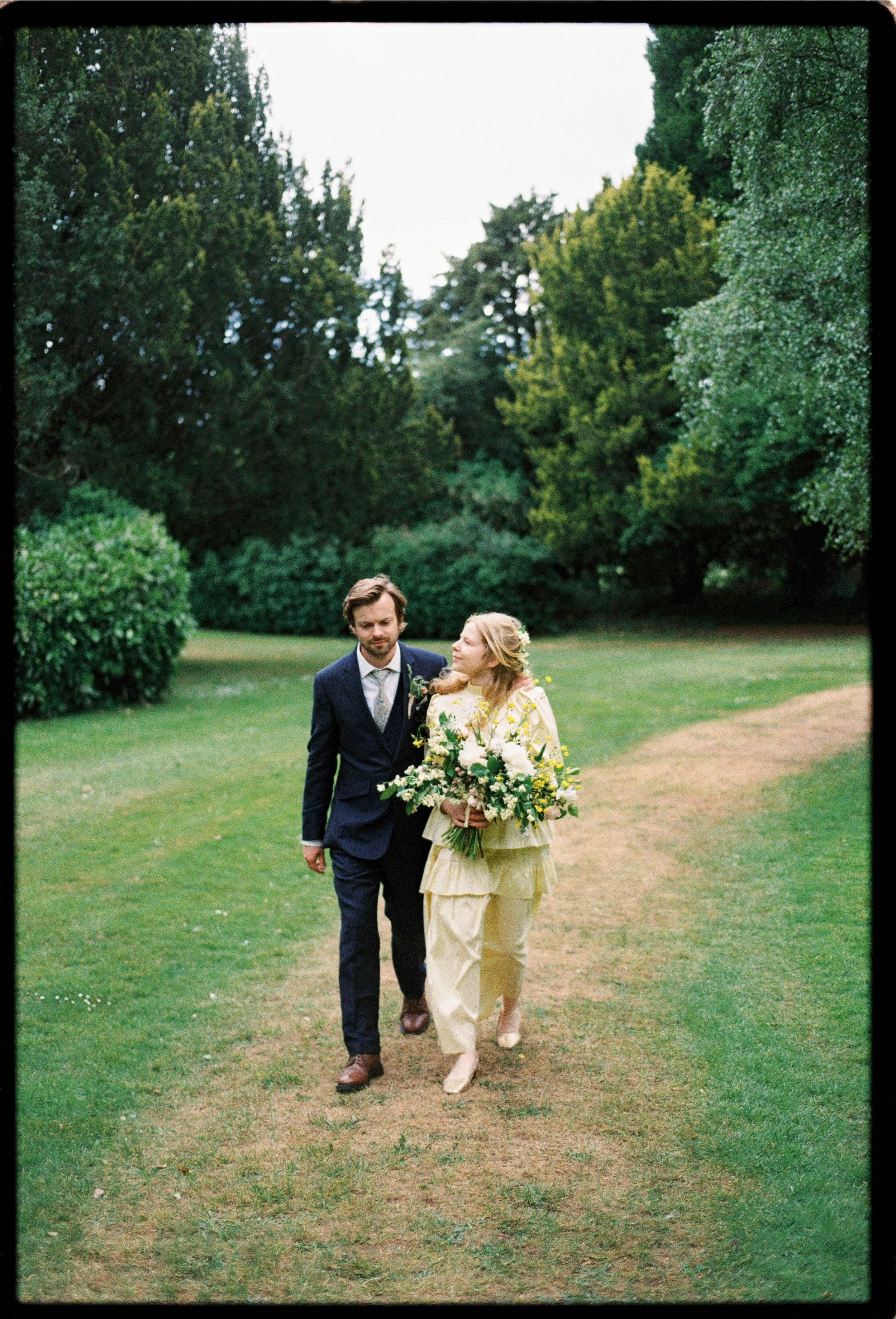 A man and woman walking on a grassy path in a park, with the woman holding a large bouquet of flowers, on a cloudy day.