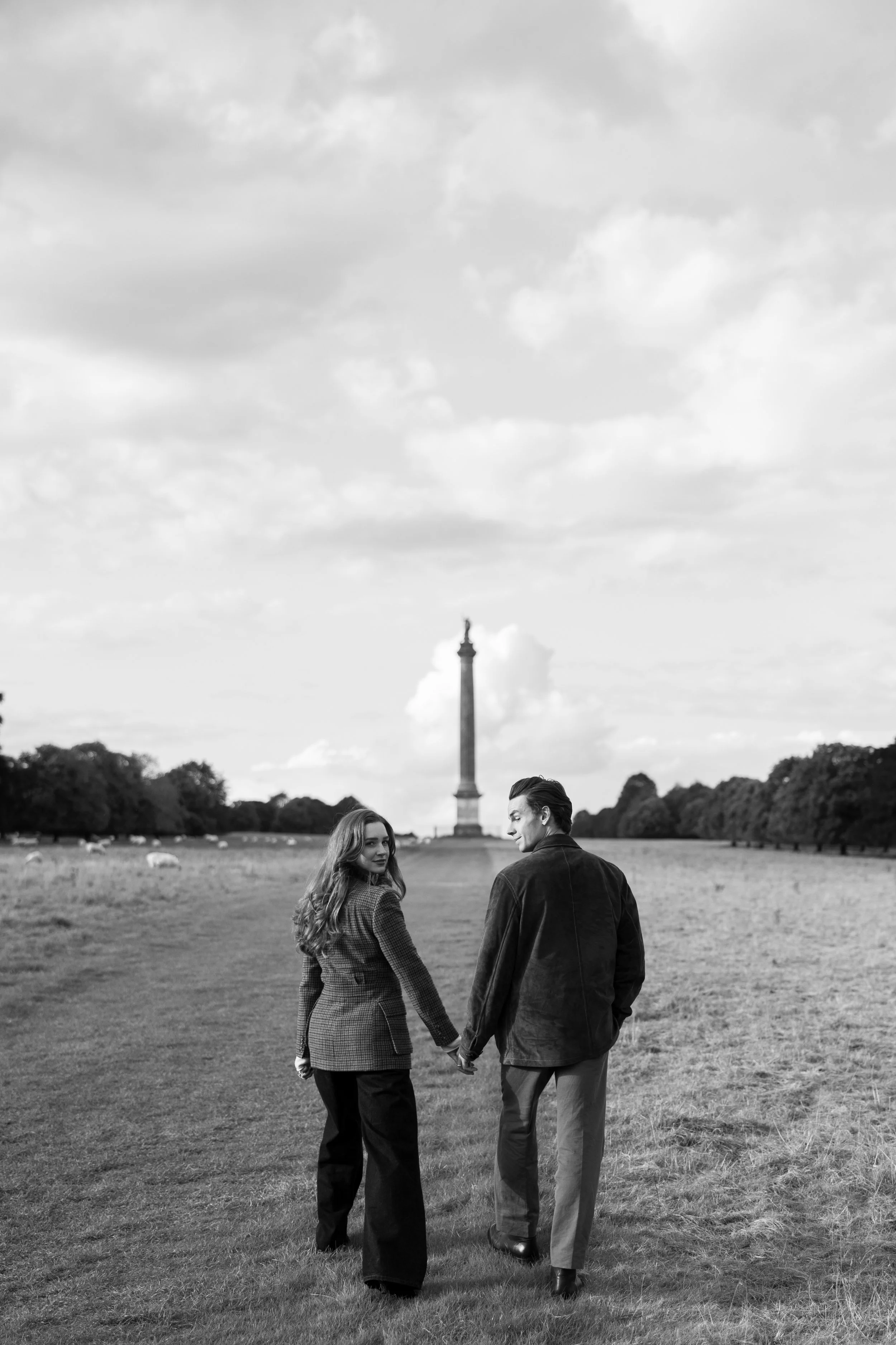 A black-and-white photo of a couple holding hands while walking in a large grassy field, with the Eiffel Tower in the background.