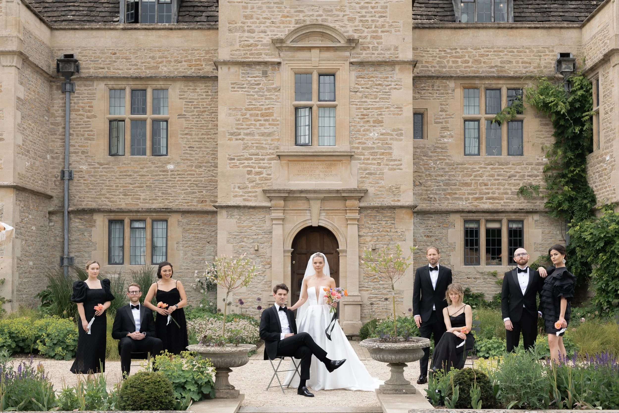 Group of wedding party members posing outside a historic stone building, with the bride in a white gown holding a bouquet, surrounded by bridesmaids and groomsmen in formal black attire.