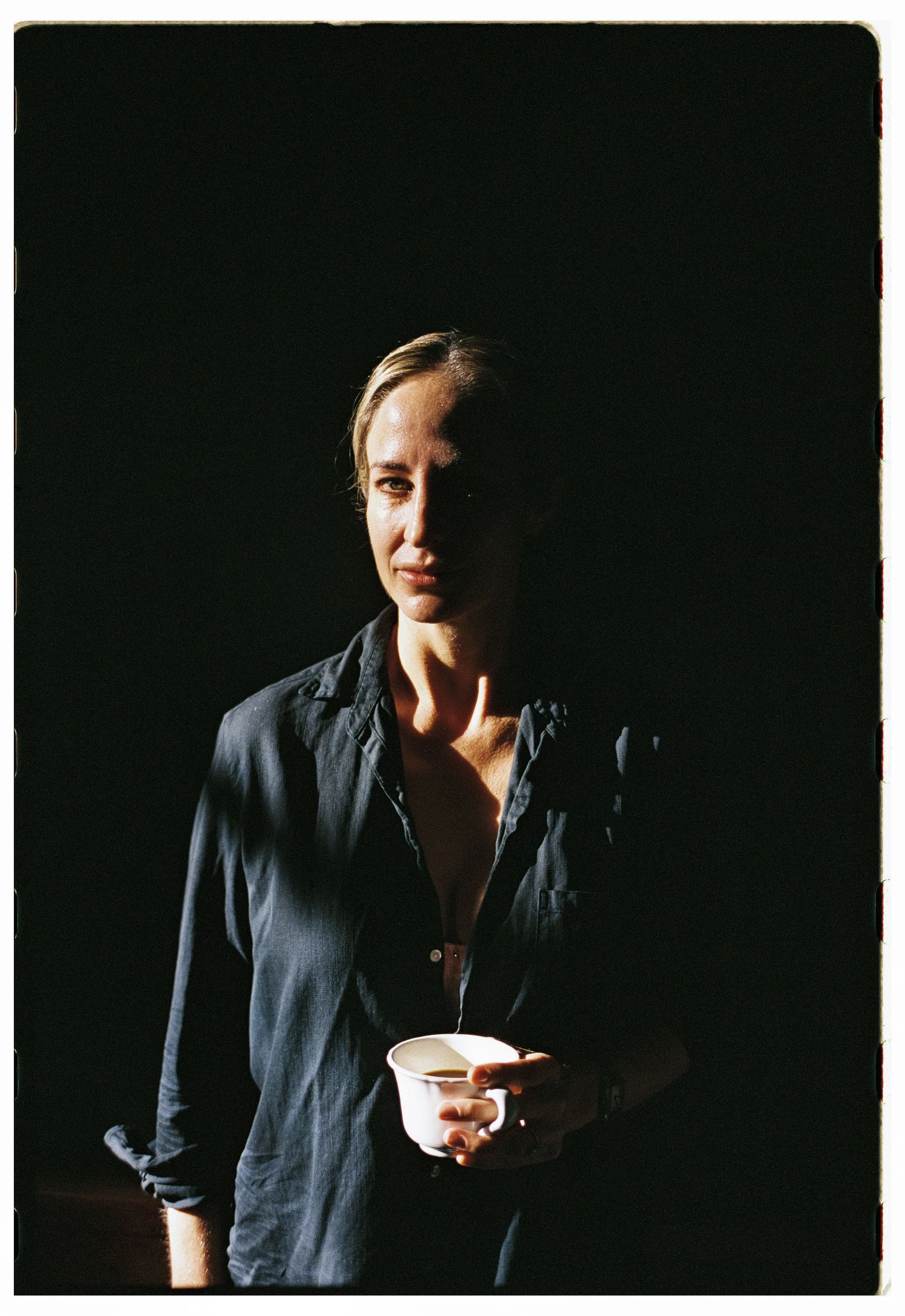 Woman holding a white mug, partially shadowed with dramatic lighting on her face, wearing a dark shirt, against a dark background.