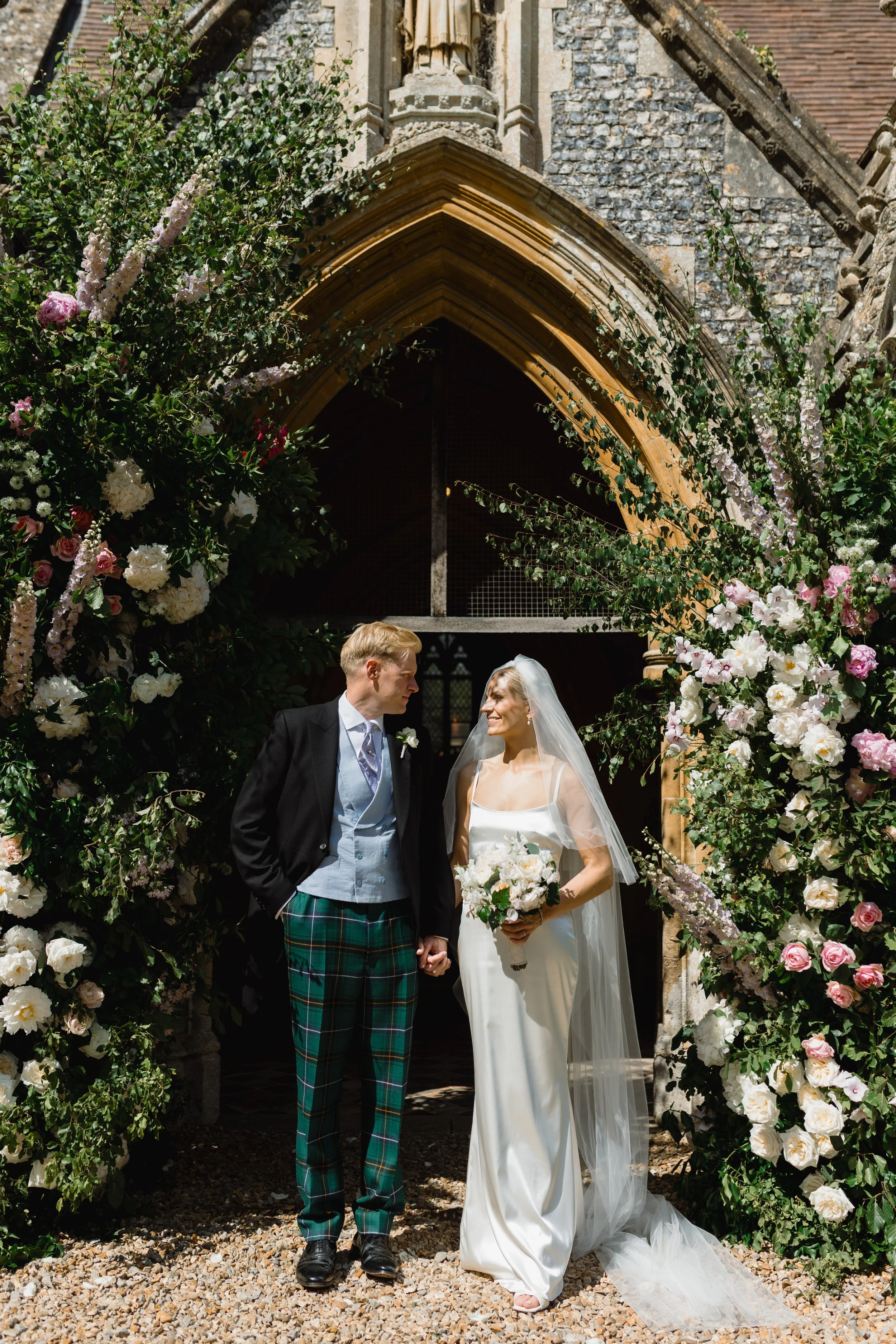 A wedding couple holding hands, standing under a floral arch outside a stone church on a sunny day.