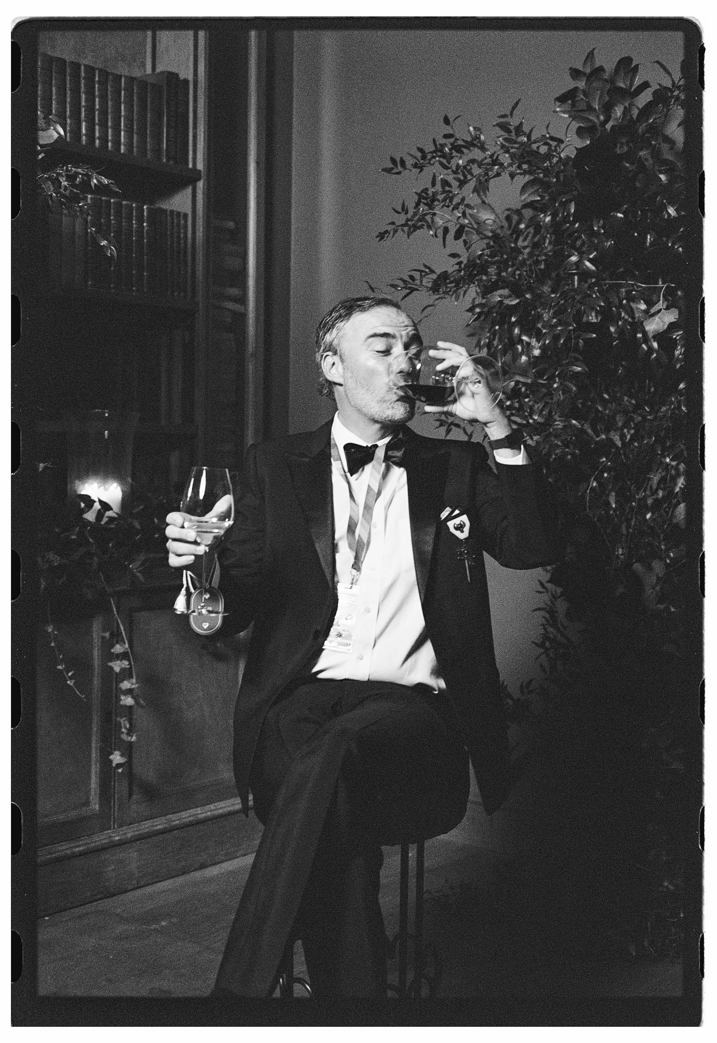 A man in a tuxedo sits on a stool, drinking wine, with a large plant and bookshelves in the background.