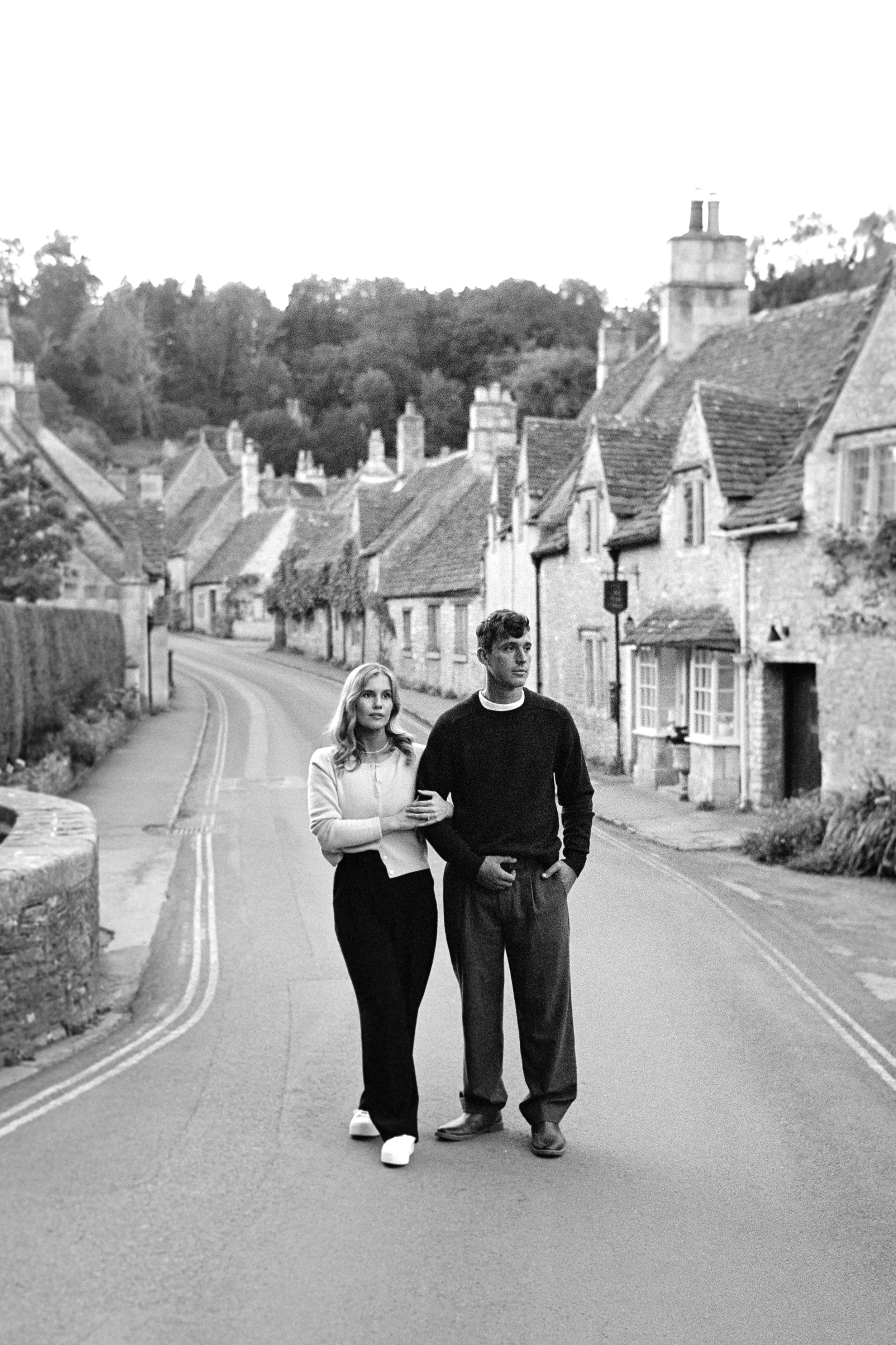 A black-and-white photo of a young man and woman walking arm in arm on a rural street lined with old houses in a quiet village.