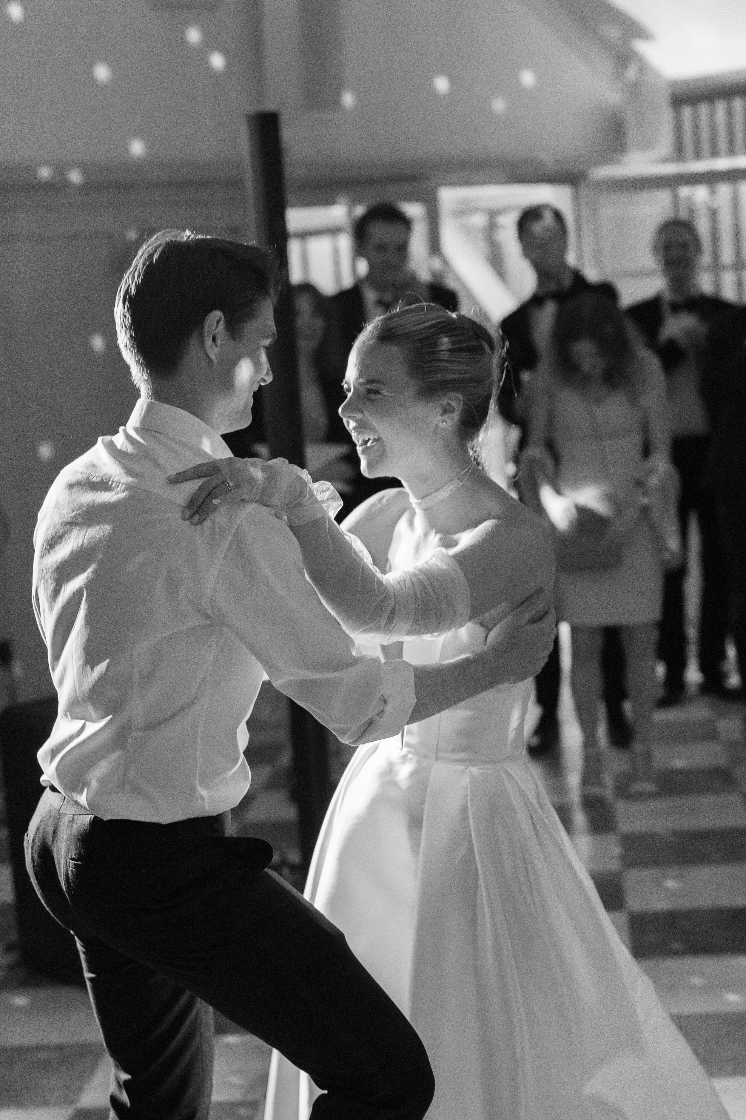 A black-and-white photo of a wedding dance with a bride and groom smiling at each other, surrounded by guests.