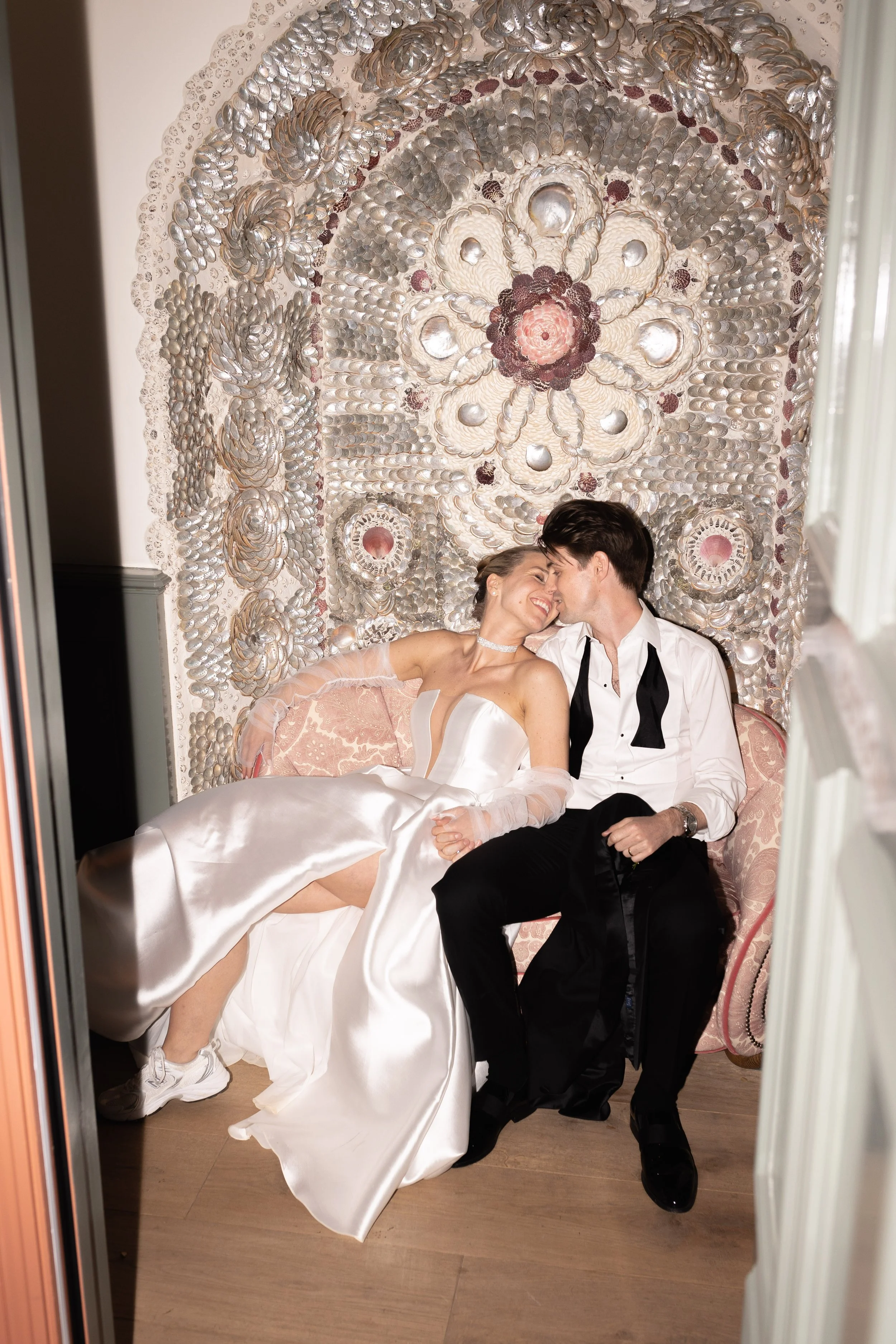 A bride and groom sitting on a pink vintage sofa with a shell and pearl decorated backdrop, smiling and leaning in for a kiss.