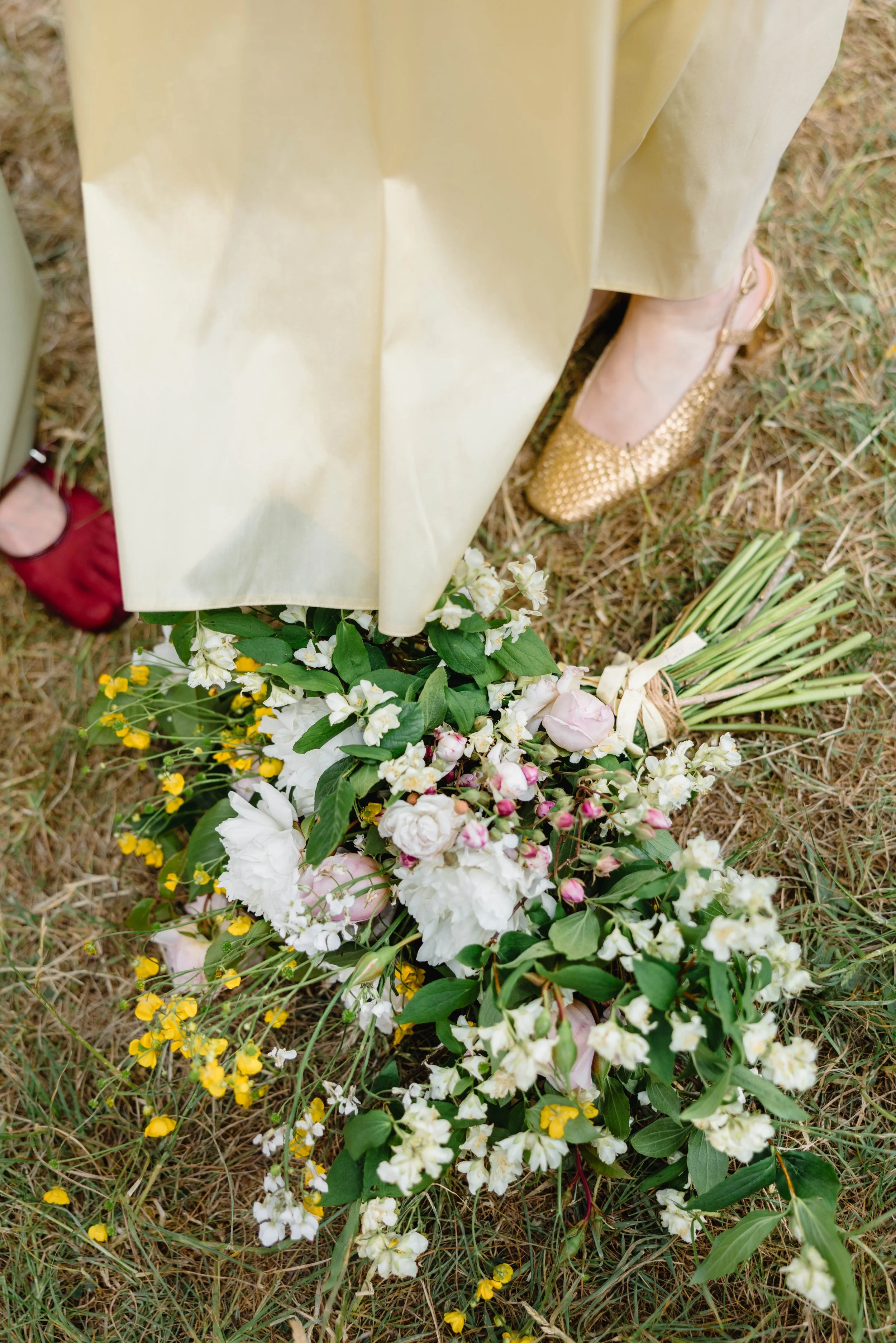 View of a person's feet standing near a bouquet of white, pink, and yellow flowers on the grass, with the person wearing beige pants and shiny gold shoes.