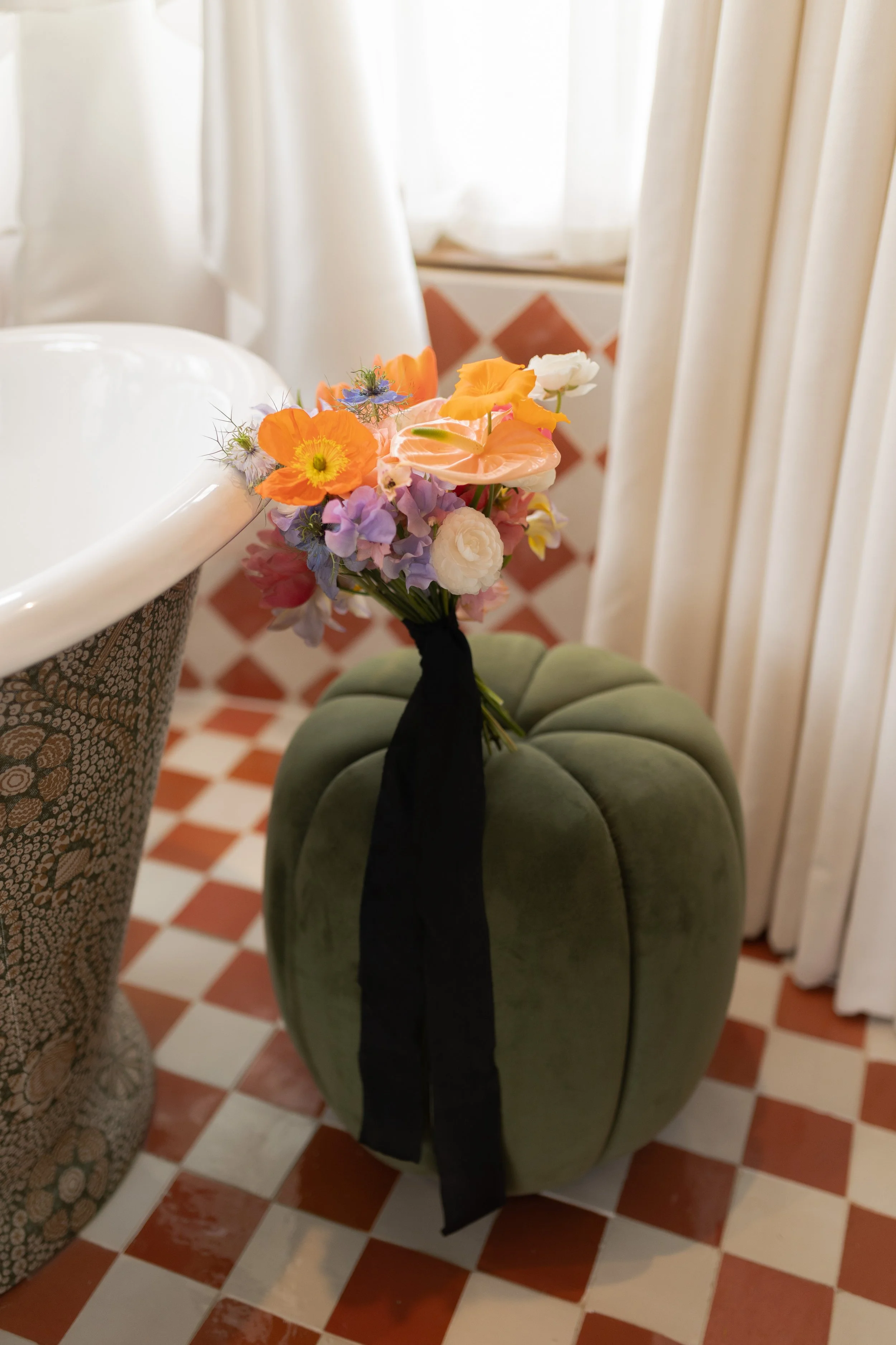 A bouquet of colorful flowers with orange, purple, pink, and white blooms, tied with a black ribbon, resting on a plush, green, pumpkin-shaped ottoman in a sunlit room.