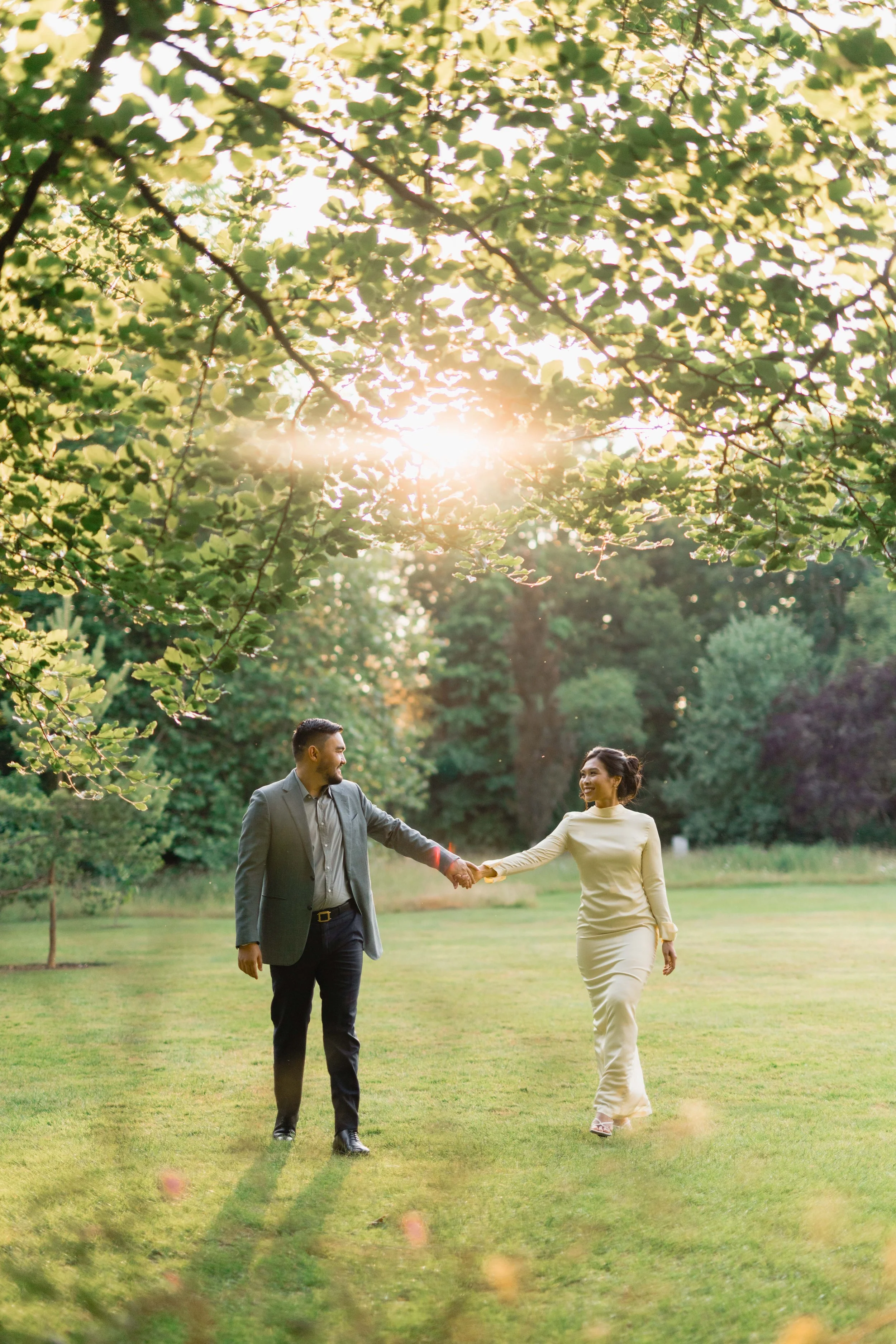 A couple holding hands and walking on a grassy field during sunset, surrounded by trees.