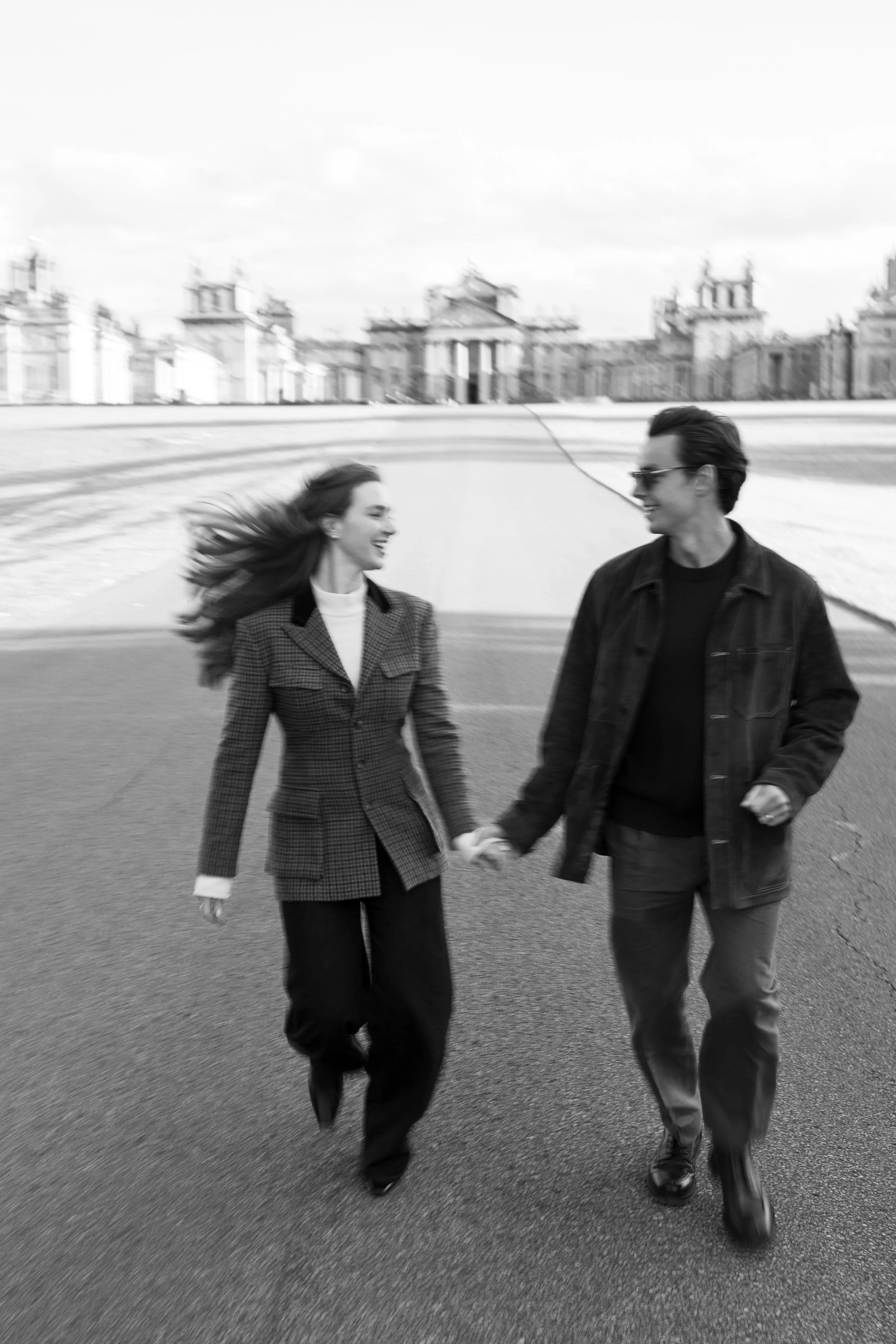 A black and white photo of a man and woman walking hand in hand outdoors, smiling at each other on a paved area with historical buildings and a cloudy sky in the background.