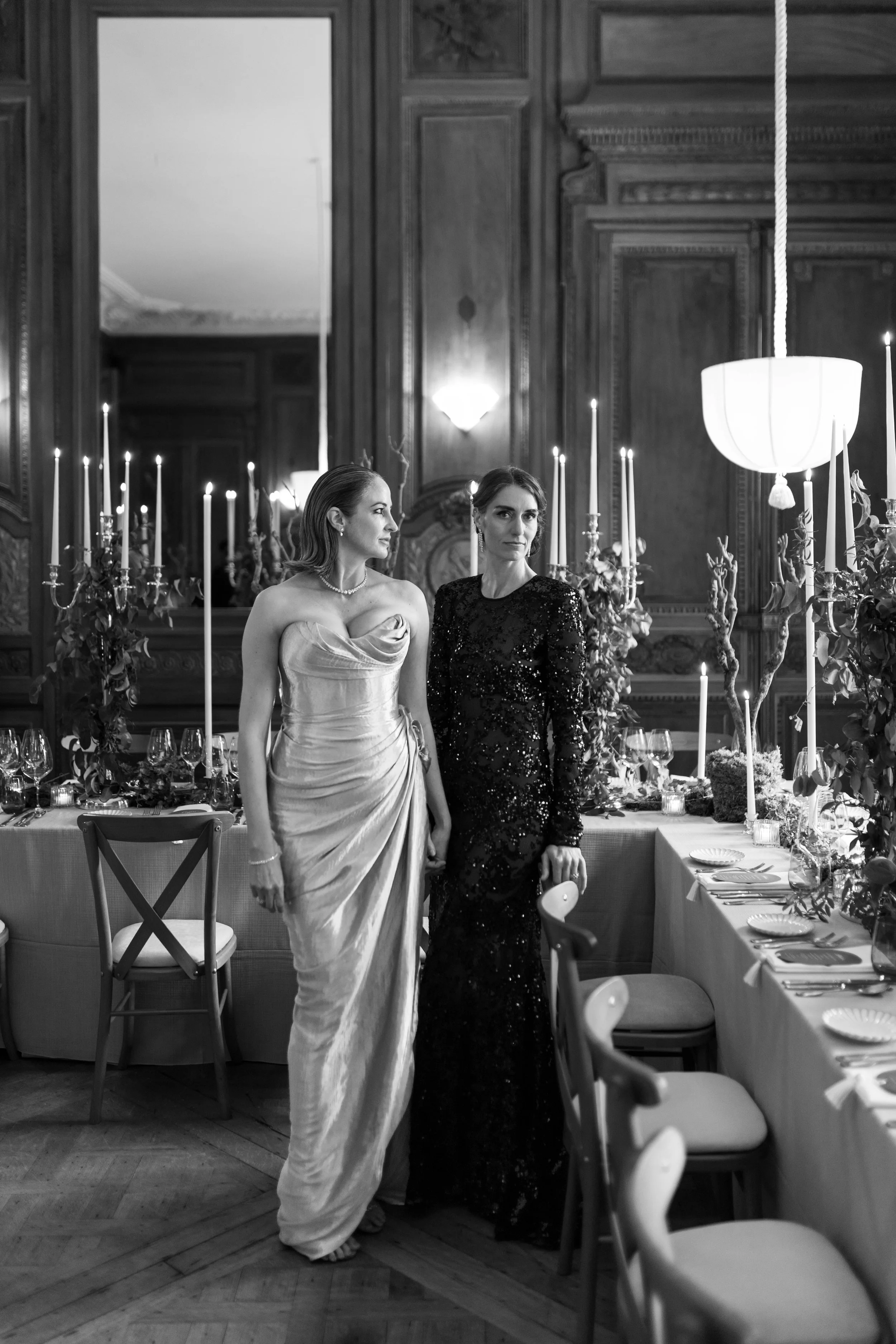 Two women in elegant evening dresses standing in a lavishly decorated dining room at Cowley Manor Experimental with candelabras, floral arrangements, and a long table set for an intimate wedding.
