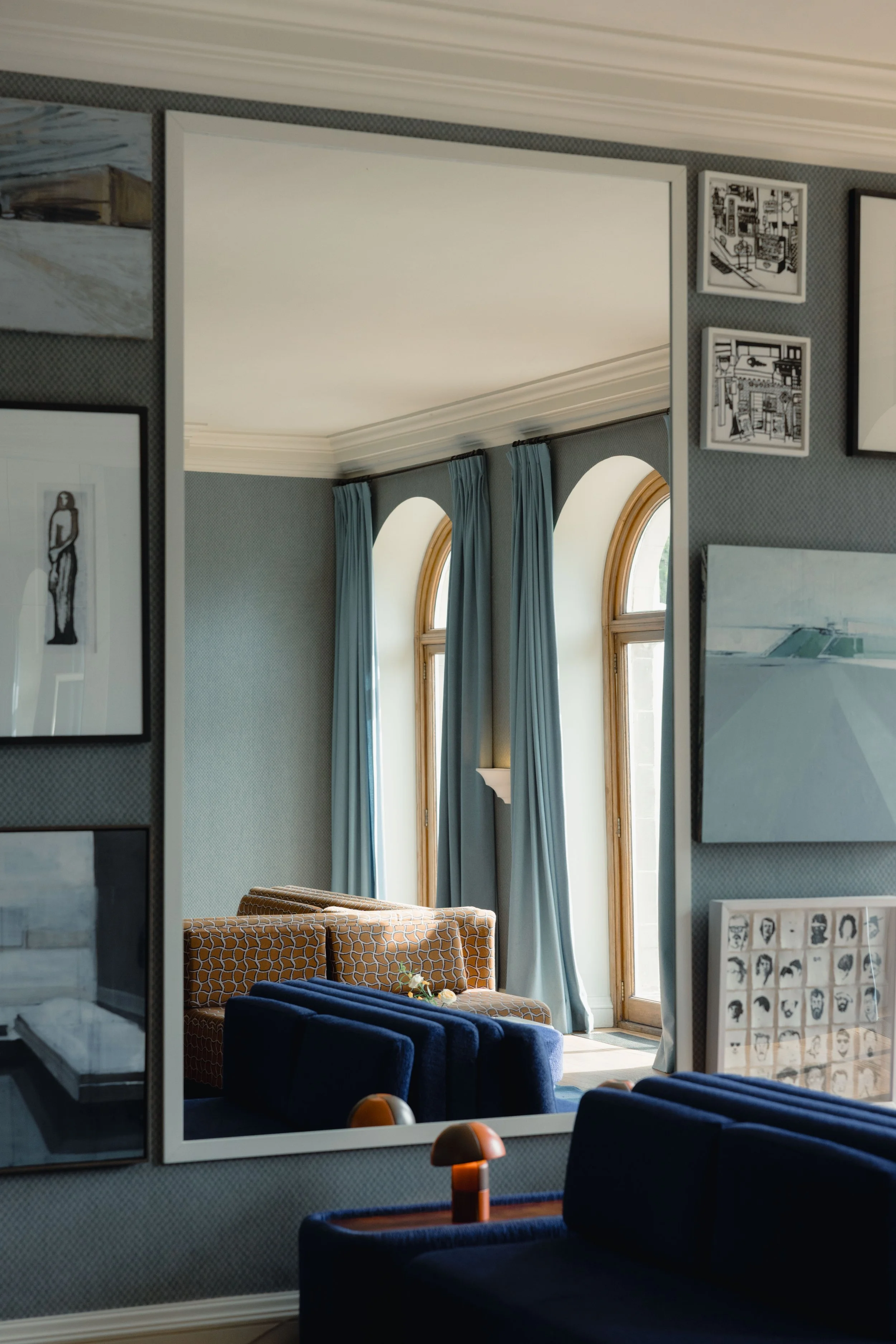 The lounge at Cowley Manor reflected in a large mirror, featuring tall arched windows with blue curtains, a brown patterned couch, and a blue velvet armchair.