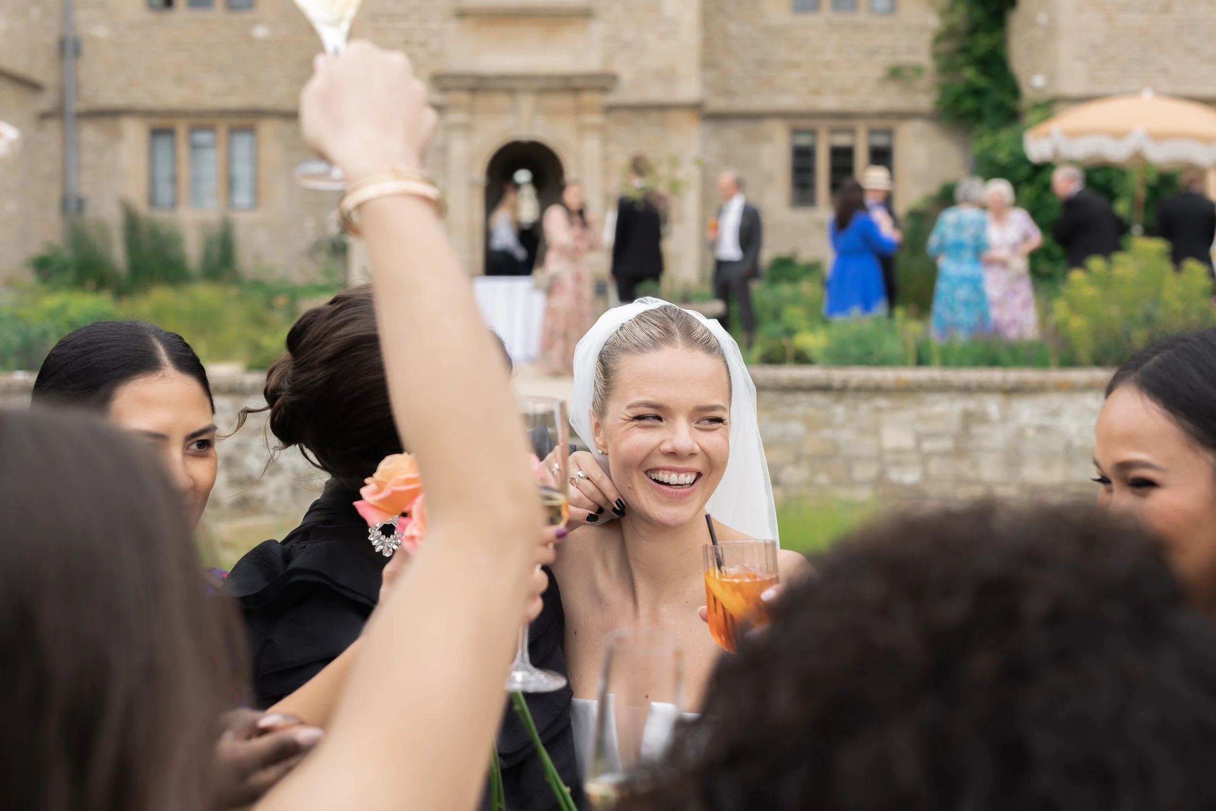A group of women celebrating outdoors at a wedding reception, with the bride smiling and holding a drink, surrounded by friends raising glasses, in front of a historic stone building with people socializing in the background.