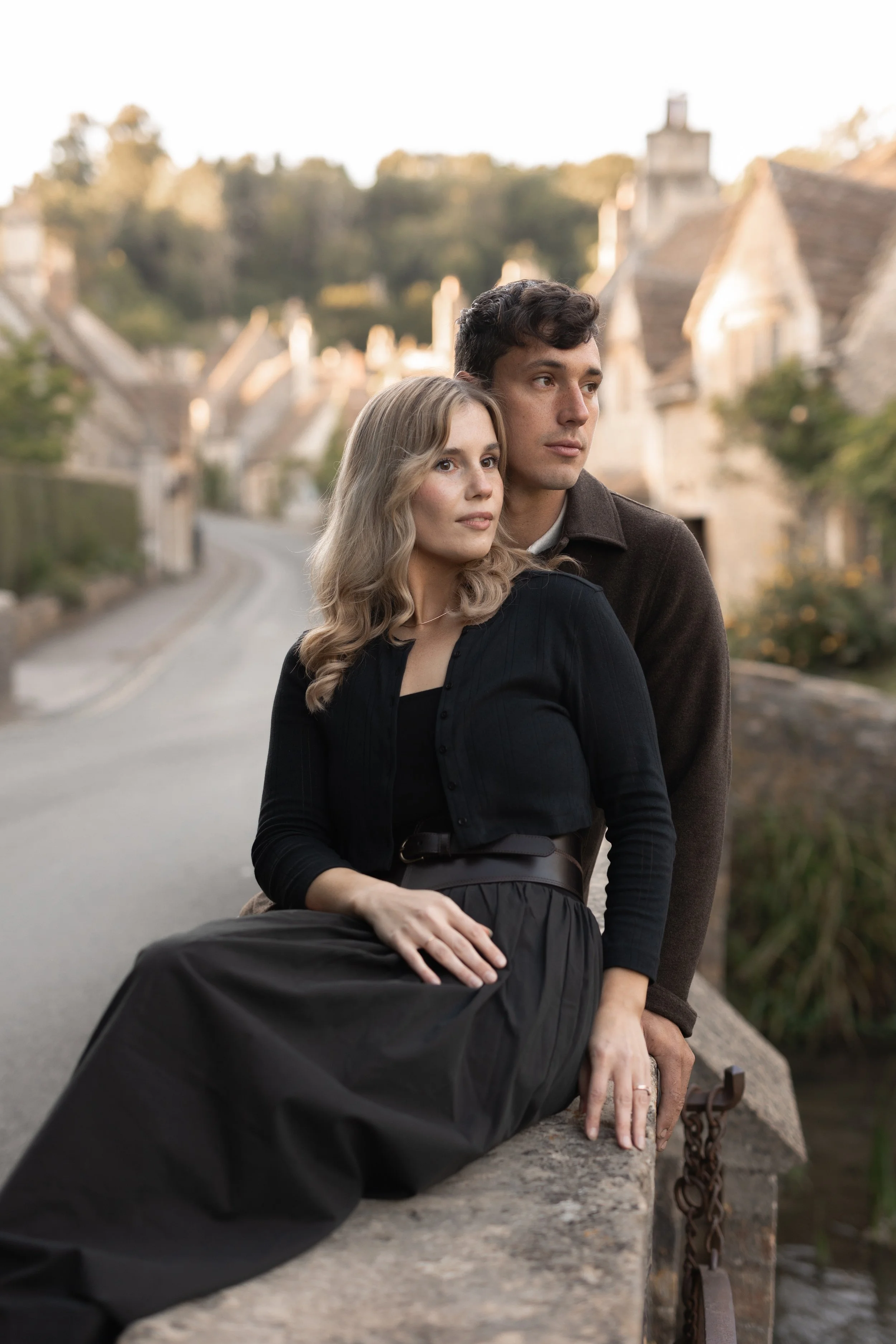 A young couple sitting on a stone wall by a river in a small European village, with old stone houses and trees in the background, during daylight.