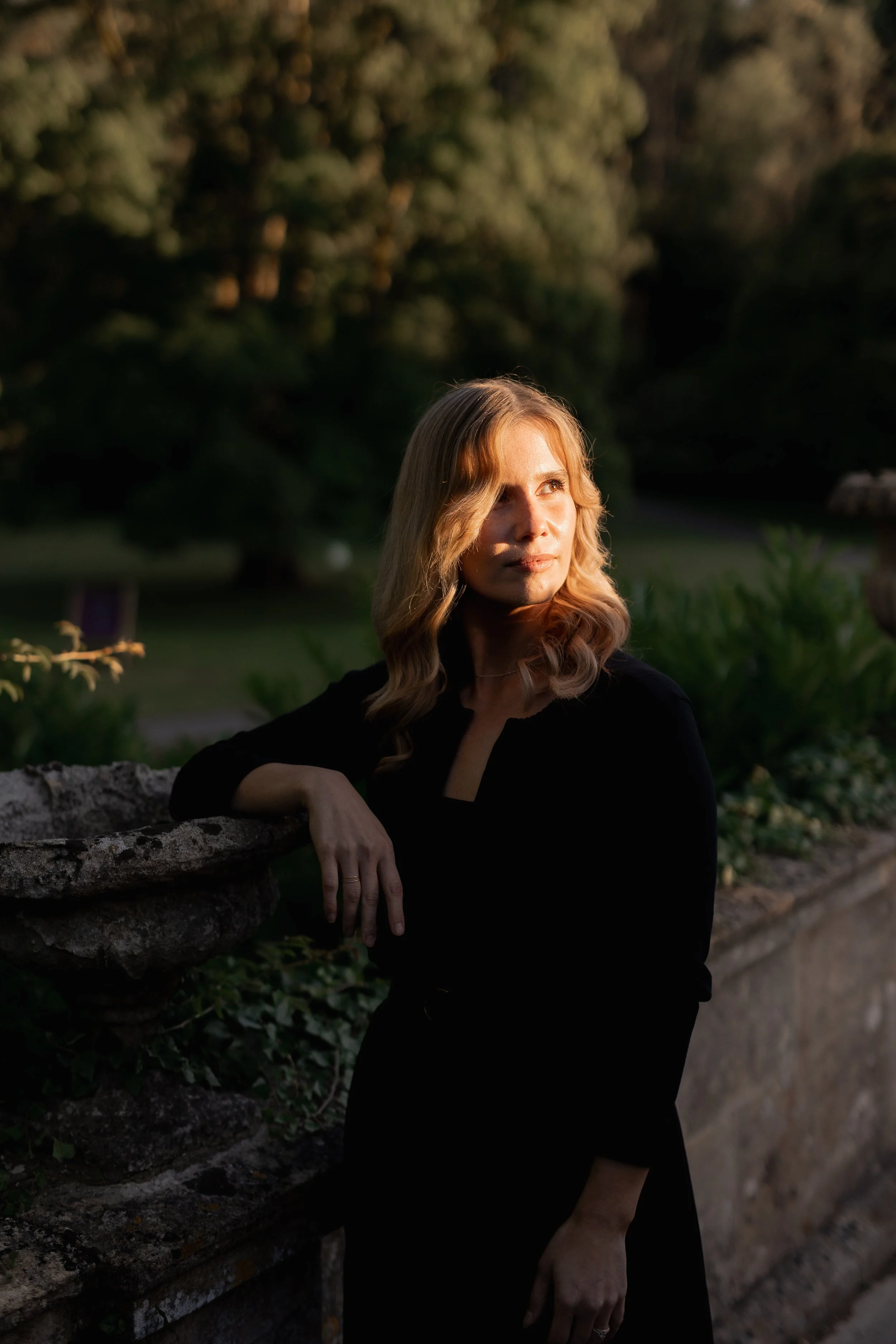 A woman with wavy blonde hair, wearing a black dress, leaning on a stone wall in a garden at sunset, looking thoughtfully into the distance.