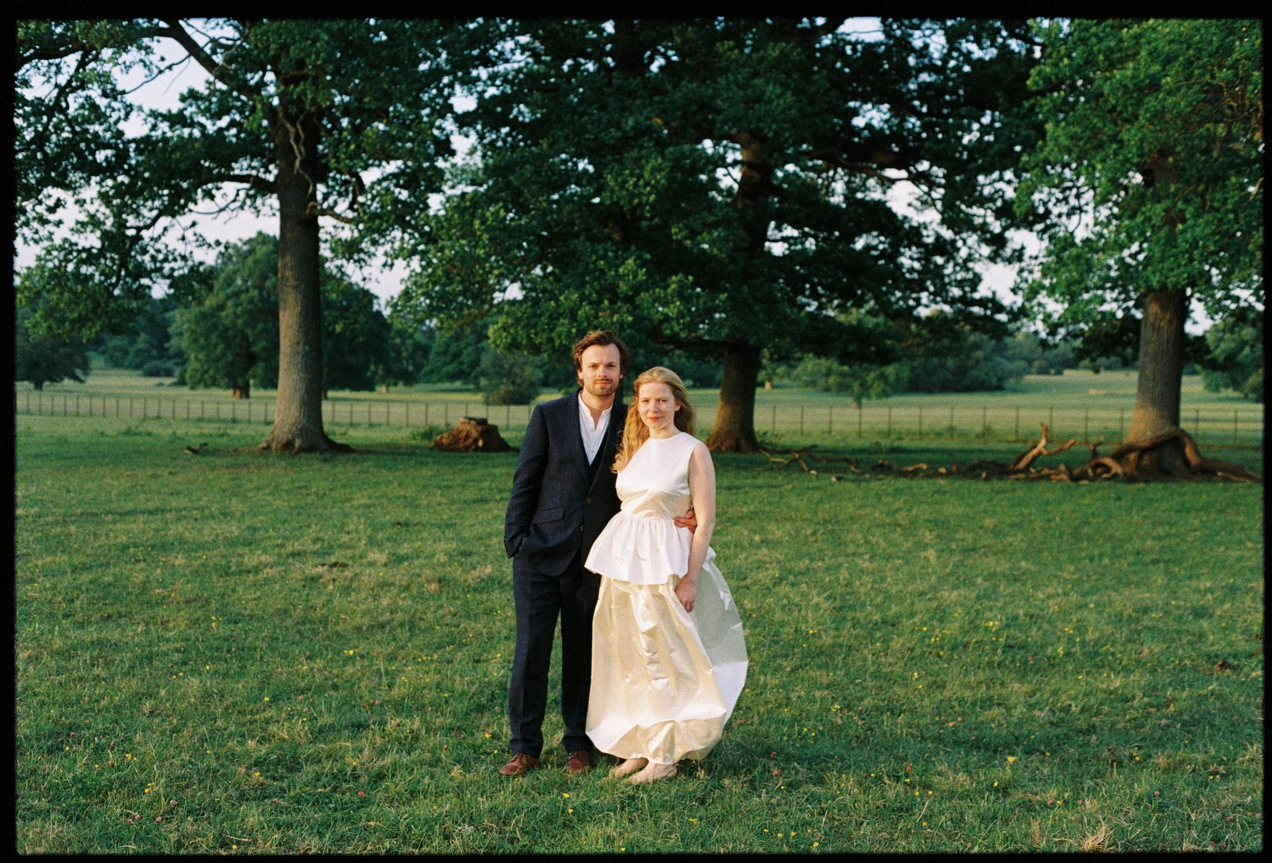 A couple standing outdoors on grass, with large trees and fields in the background. The man is wearing a dark suit, and the woman is wearing a white dress.