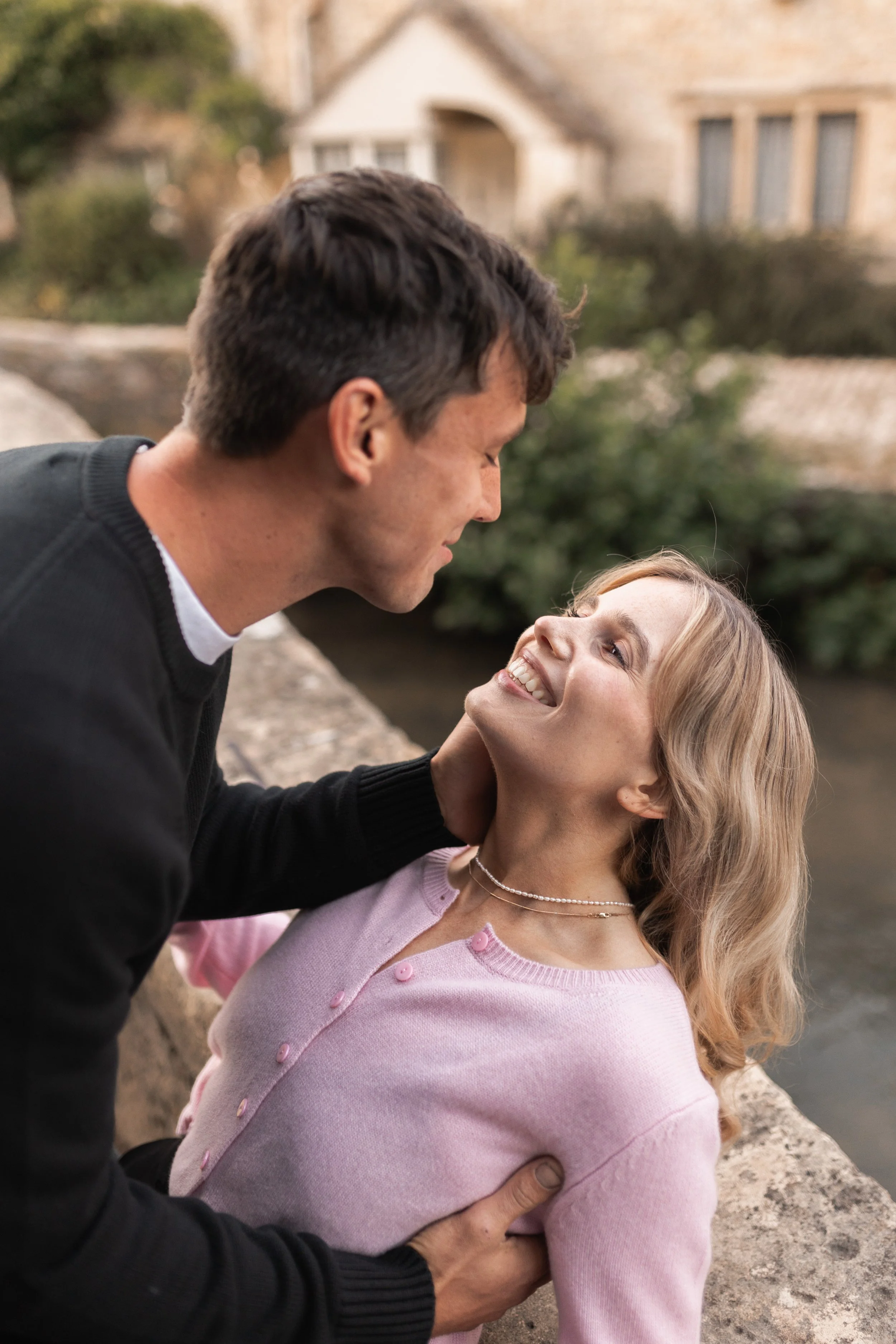 A man and woman sharing an intimate moment outdoors near a river, with the man holding the woman around her waist and her smiling up at him.