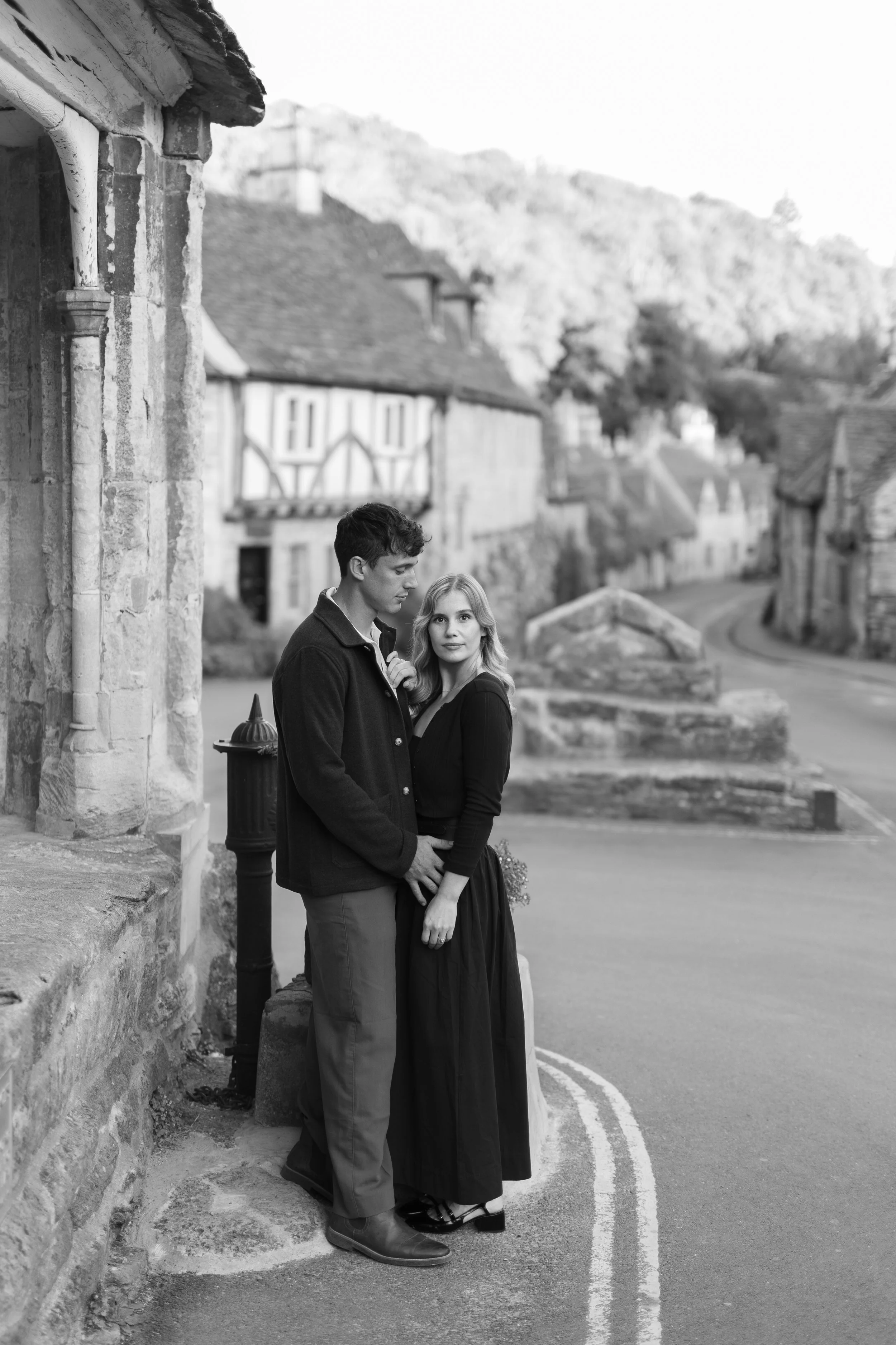 A black and white photo of a young couple standing on a street corner in a small town with historic buildings and a hillside in the background.