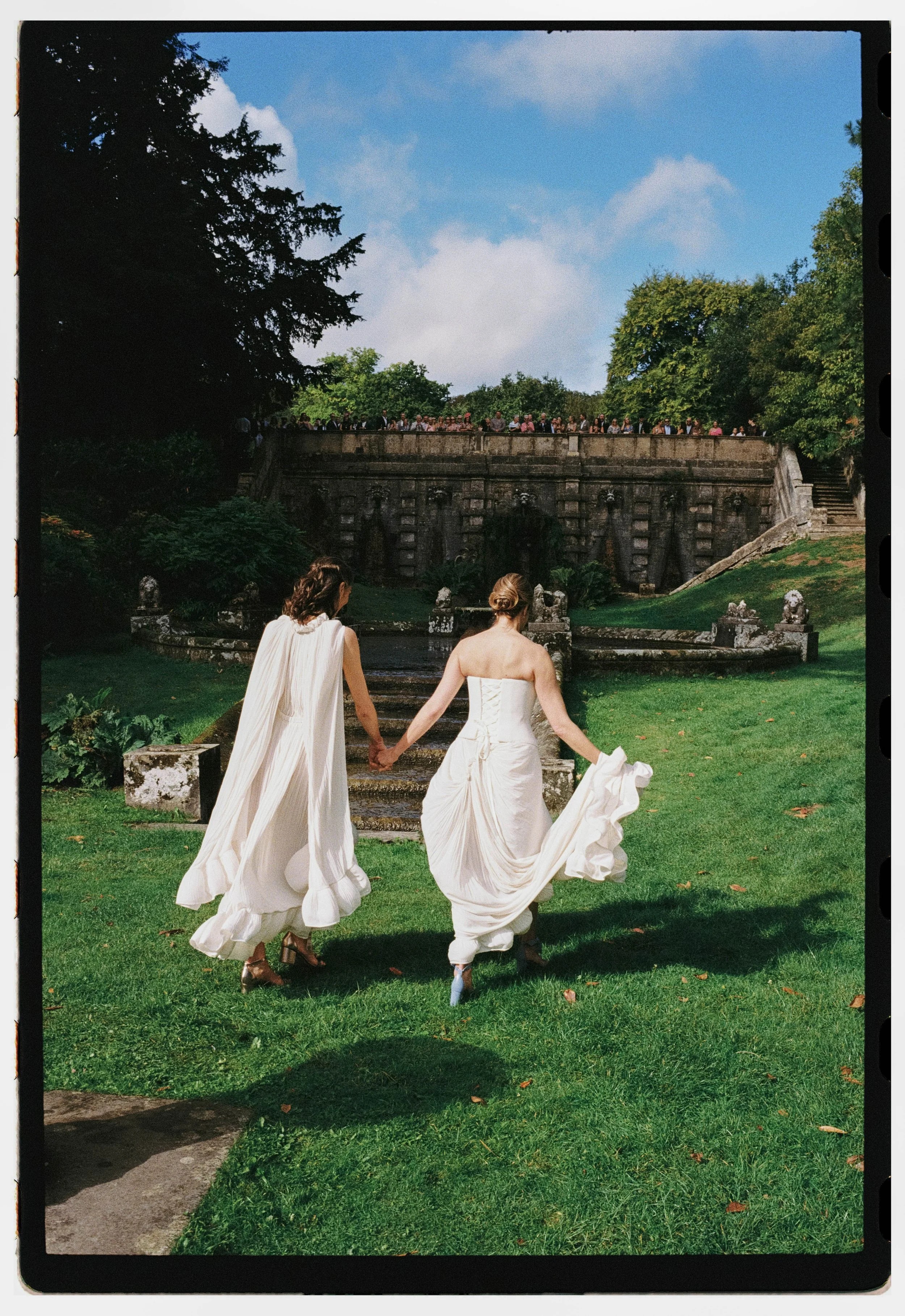 Two brides in white dresses walking hand-in-hand on a grassy area in front of a stone fountain and steps, with a crowd of people on a bridge and trees in the background under a blue sky with clouds.