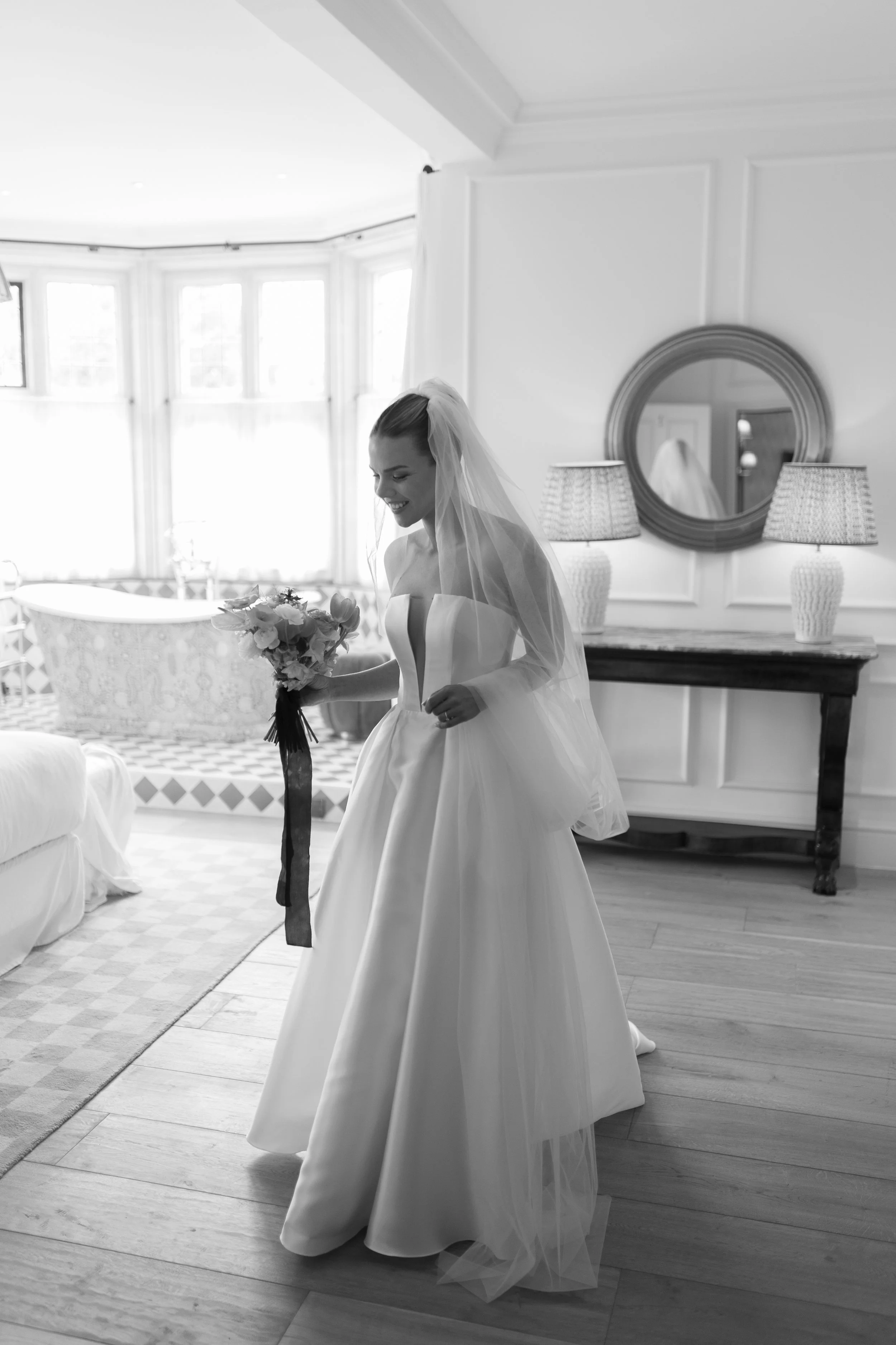 A bride in a strapless wedding gown and veil, holding a bouquet, smiling in a well-lit room with windows, a mirror, and decorative lamps.