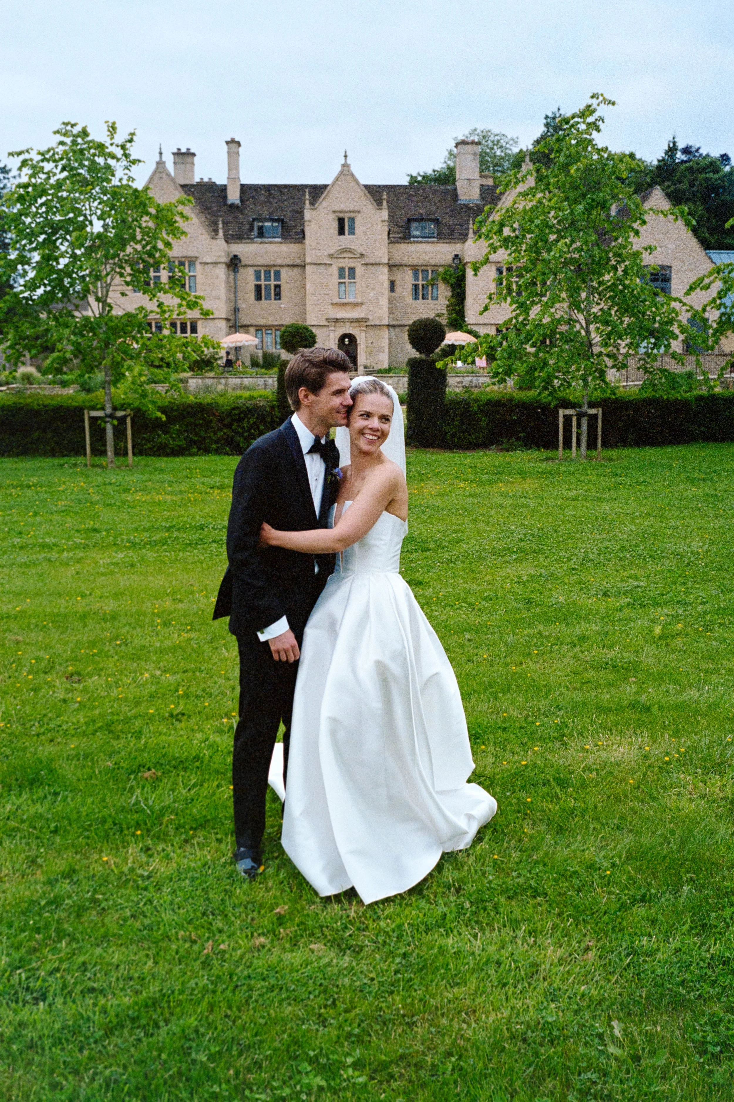 A newlywed couple in wedding attire stands on a lush green lawn, smiling and hugging with a large historic mansion in the background.