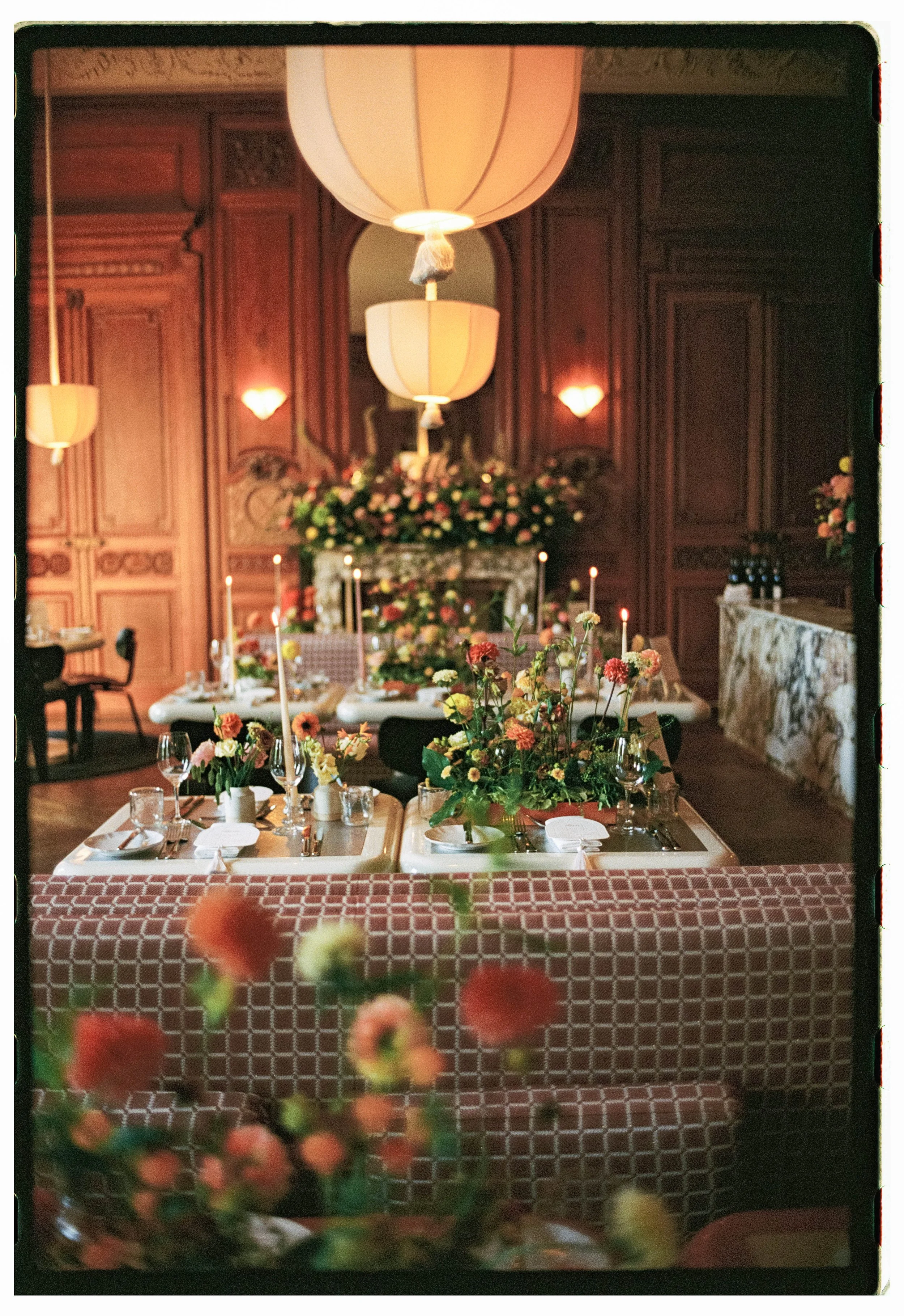 Elegant dining room with floral centerpieces, candles, and set tables, decorated for a special event in a wood-paneled space with large hanging lamps.