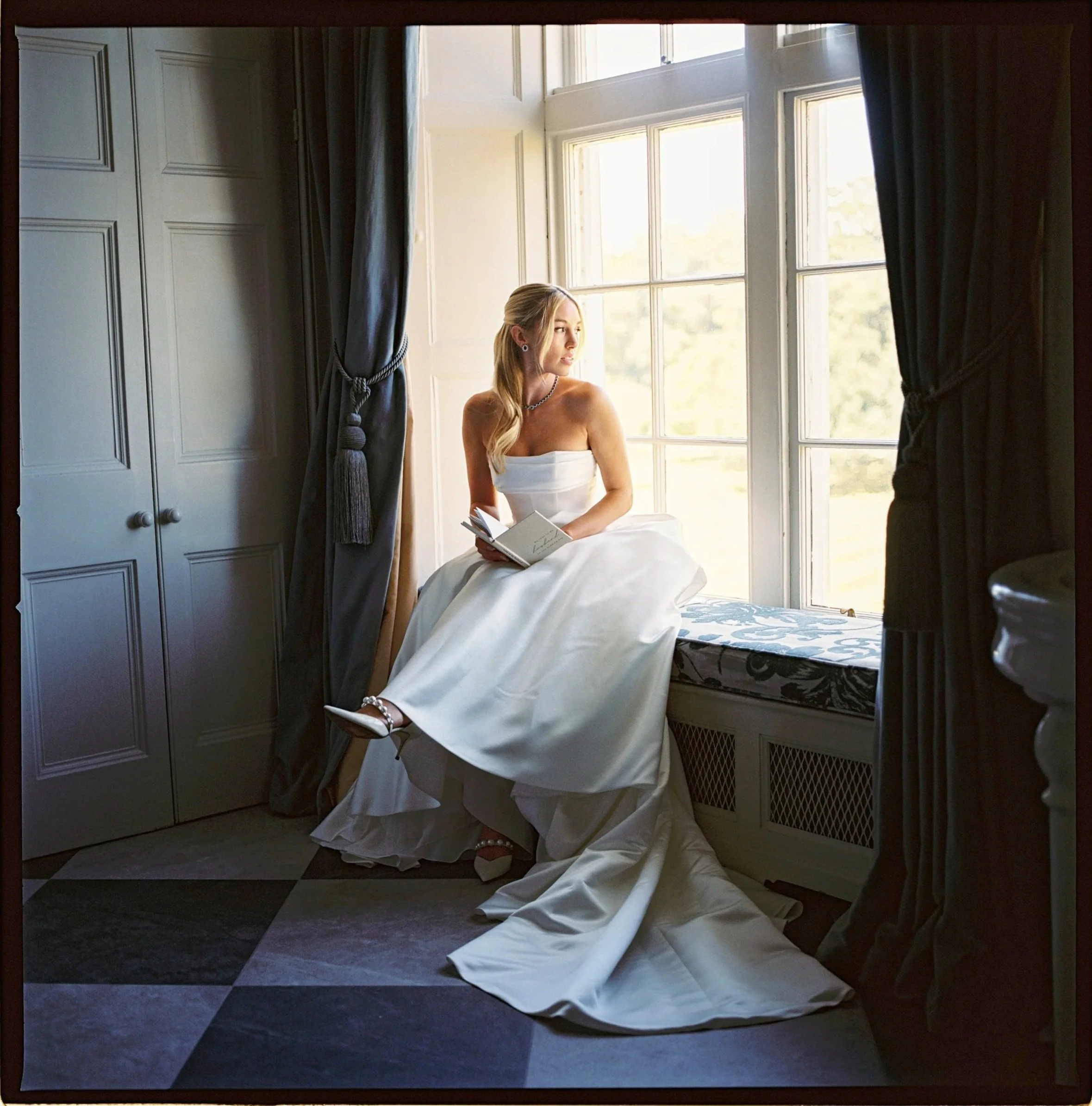 A bride sitting on a window seat, wearing a white wedding dress and pearl shoes, looking thoughtfully out the window while holding a book.