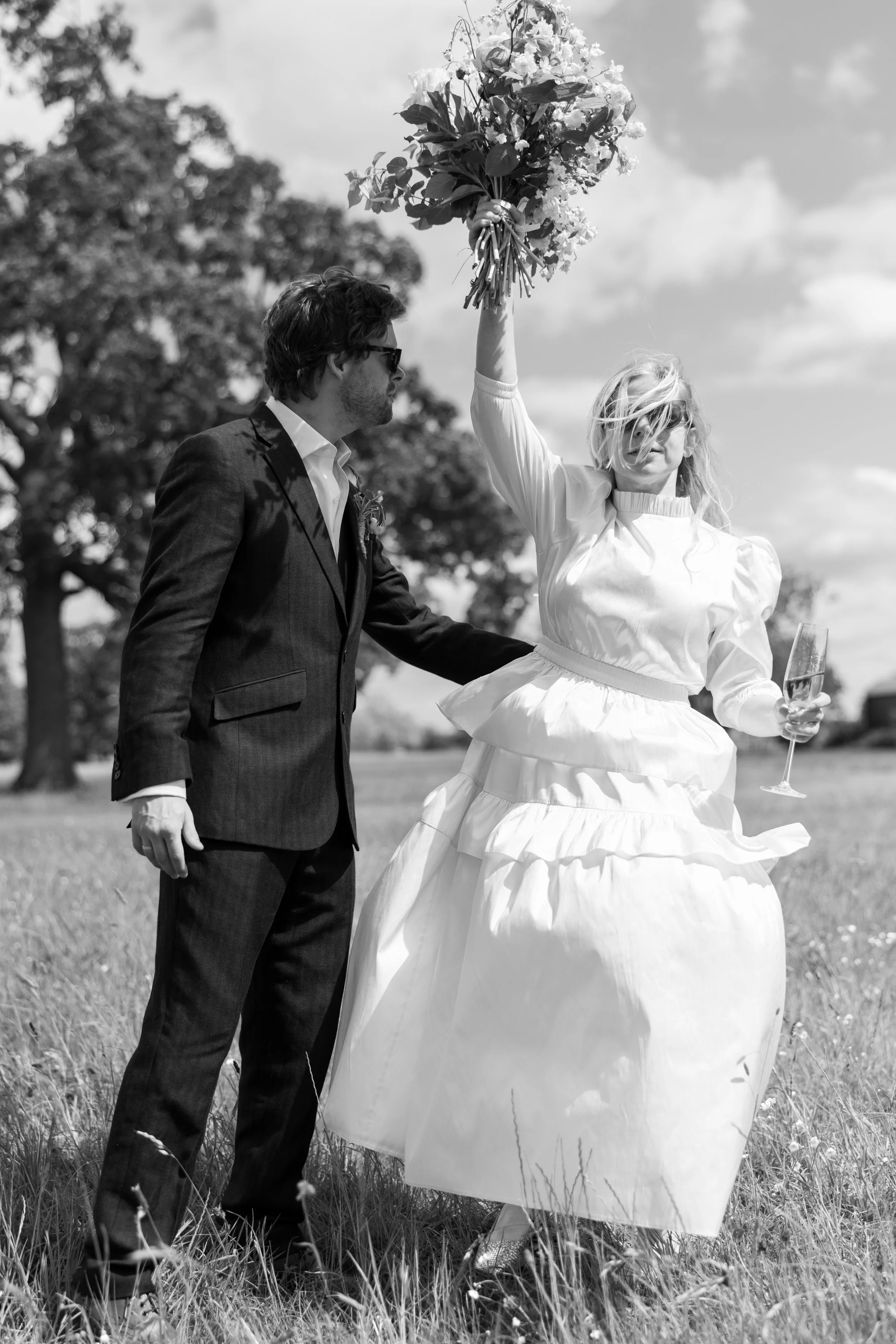A woman in a wedding dress holding a bouquet of flowers and a glass of wine, raising the bouquet in the air, with a man in a suit beside her, in an outdoor setting with trees.