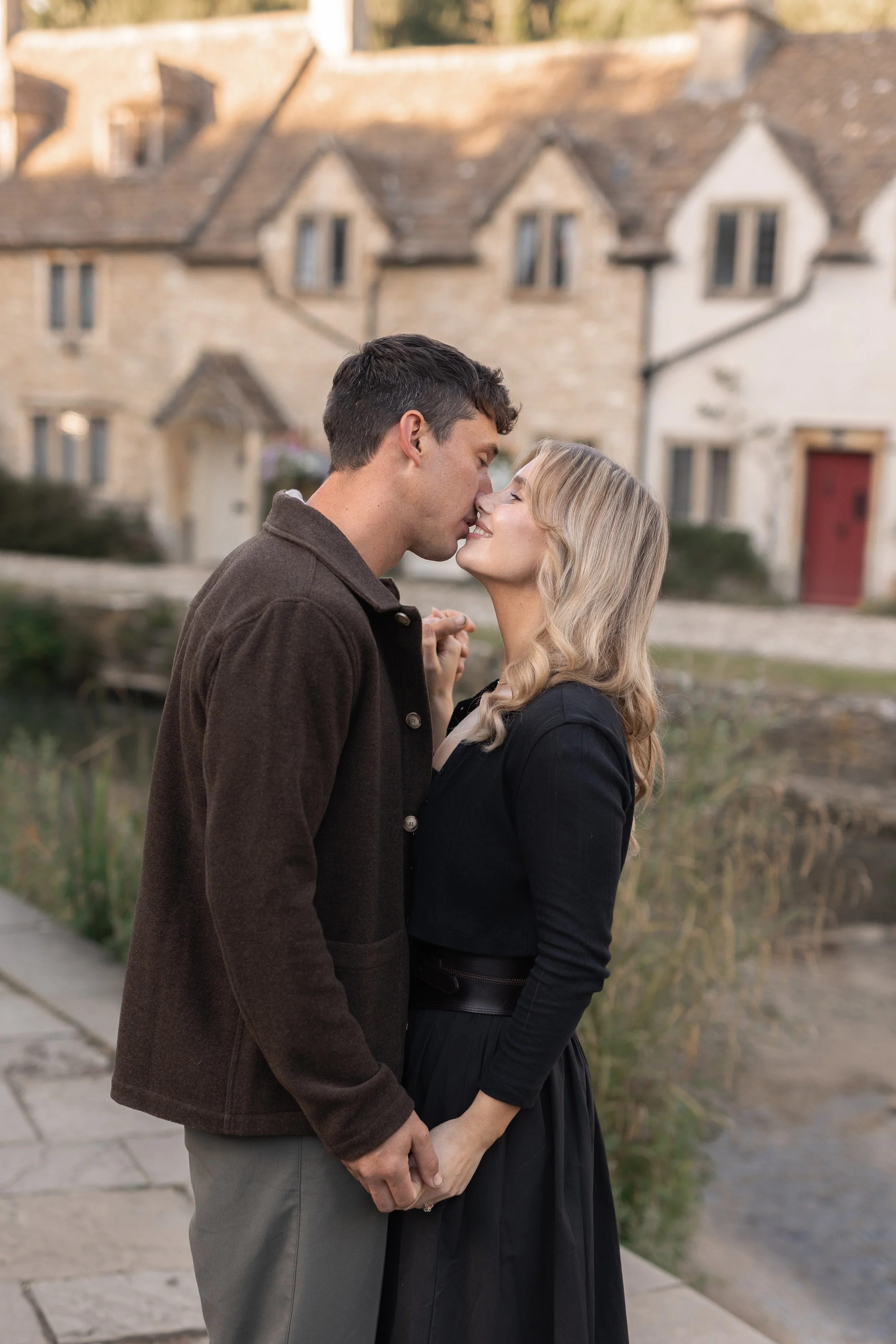 A couple holding hands and about to kiss outdoors near a pond with stone houses in the background.