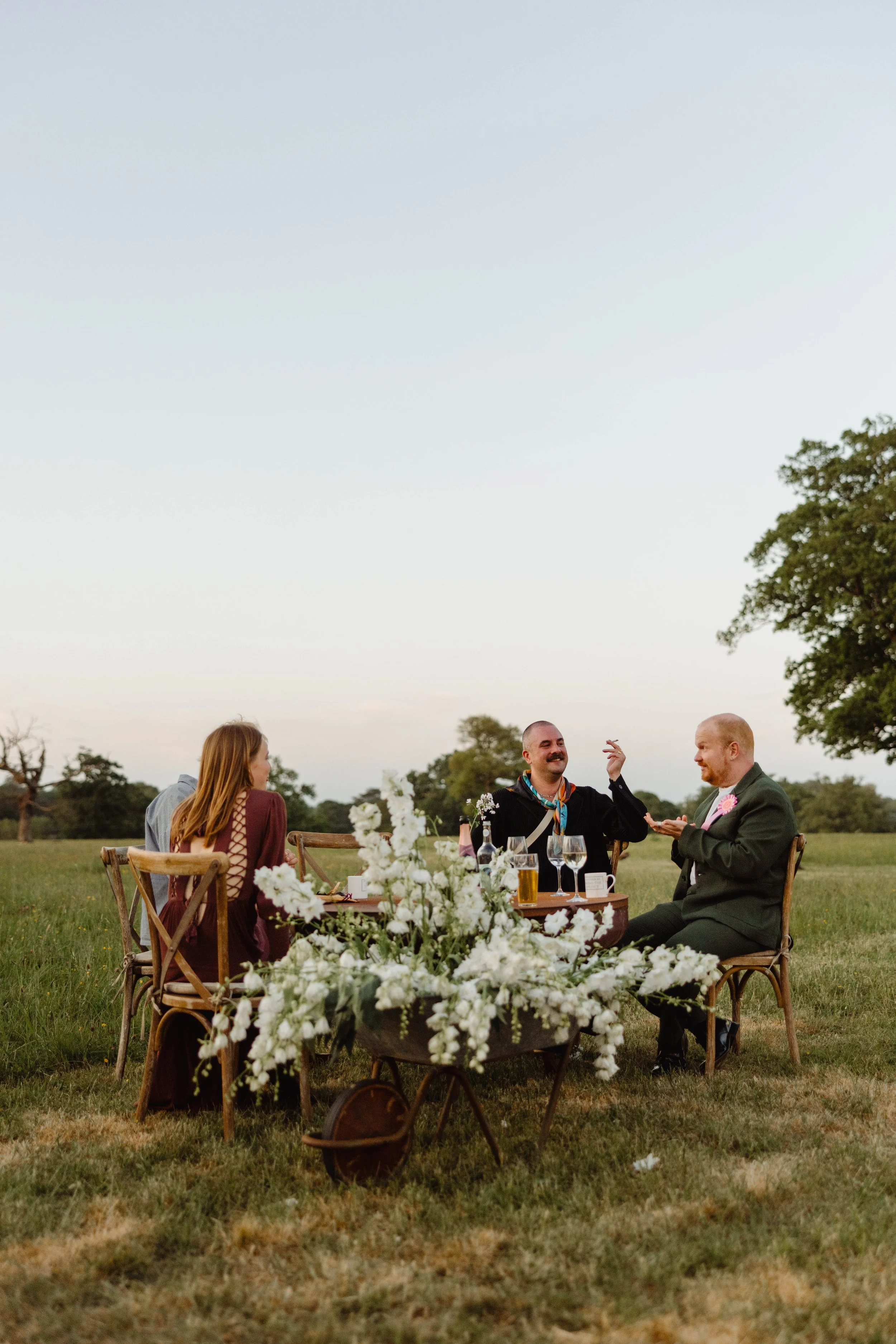 Group of four people sitting at a table outdoors during a celebration, with trees and open sky in the background. They are smiling and engaged in conversation, with drinks and a floral centerpiece on the table.