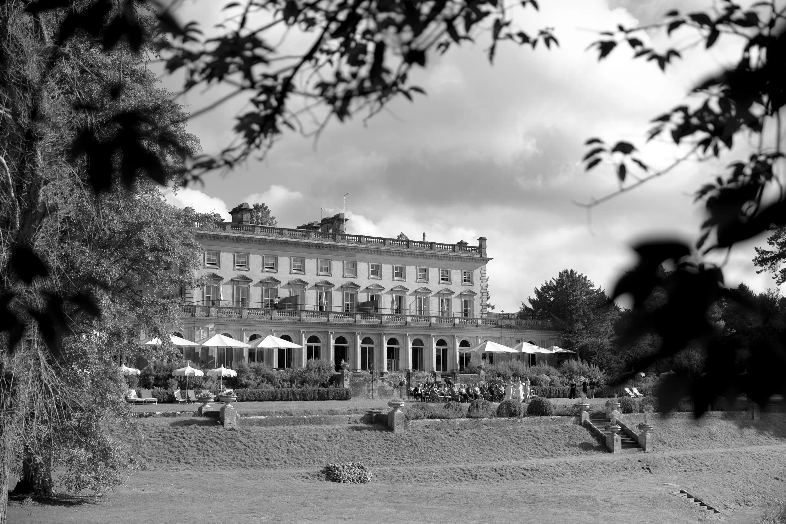 A black-and-white photo of Cowley Manor a large, elegant historic building with multiple windows, balconies, and columns, situated behind a landscaped garden with umbrellas and people gathered outdoors, framed by trees and branches in the foreground.