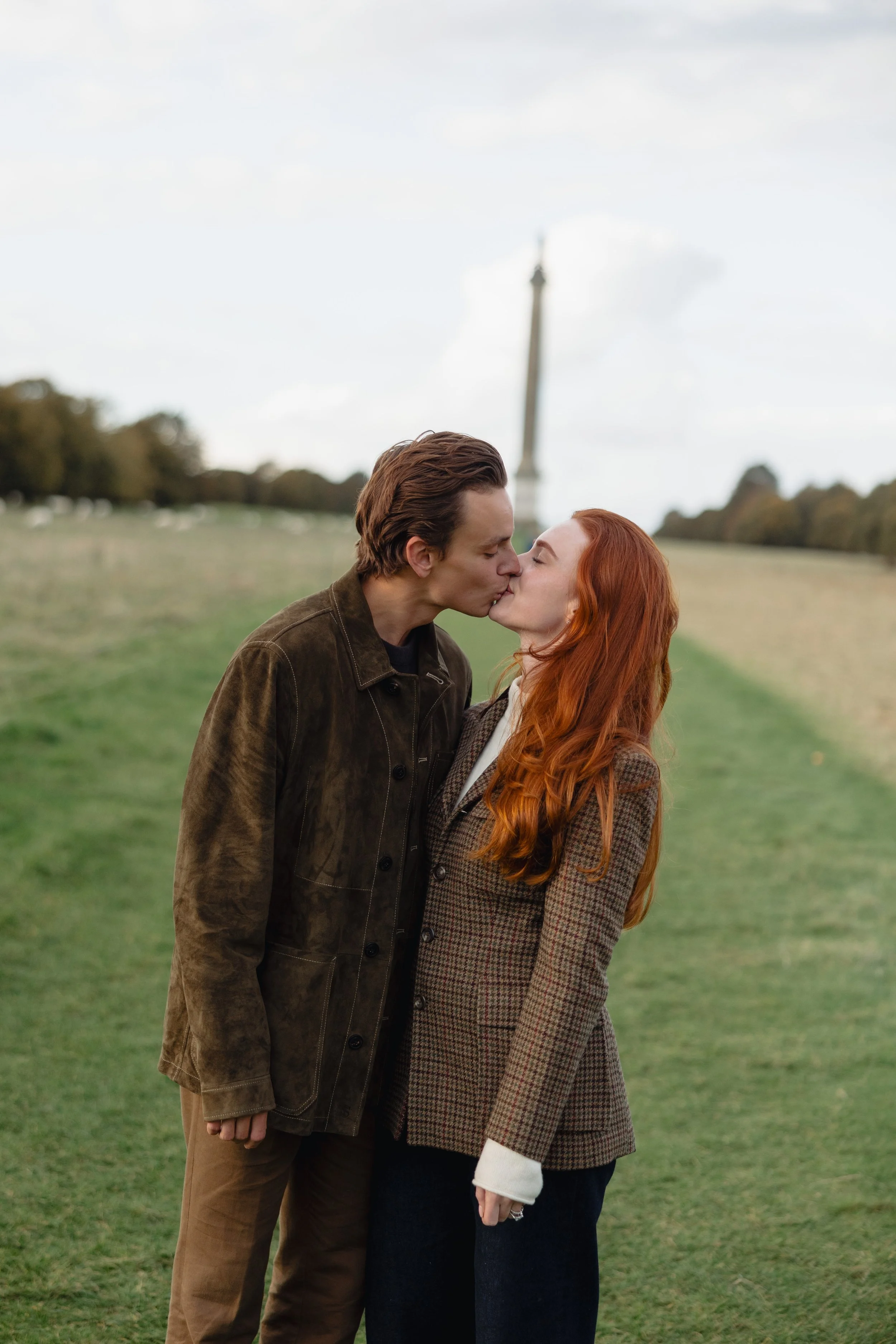 A couple with brown and red hair kissing in a park with the Eiffel Tower in the background.