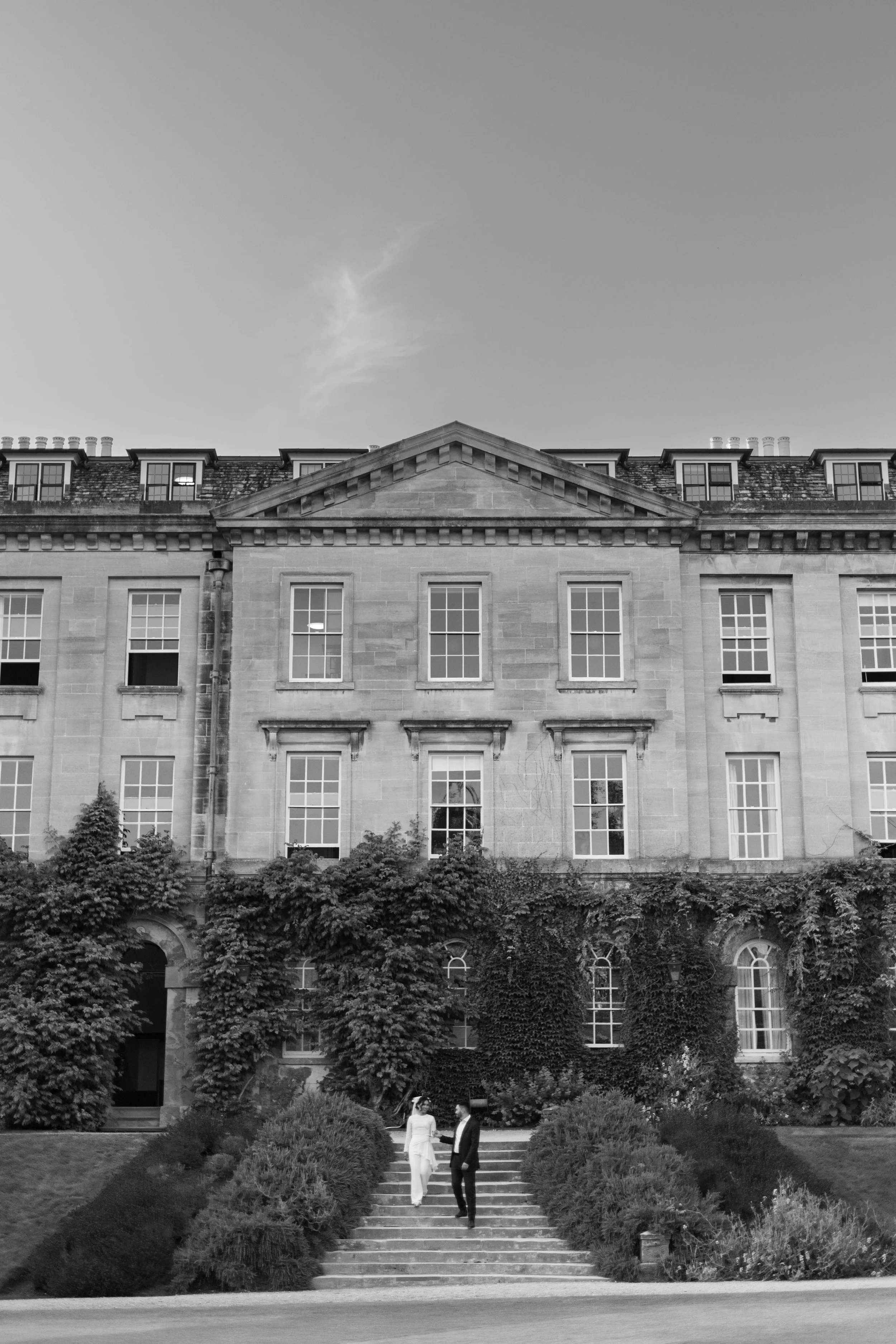 Black-and-white photo of a large multi-story stone building with ivy on the facade, multiple windows, and a triangular pediment at the roof peak. Two people are walking down the steps in front, one in light clothing and the other in dark clothing.