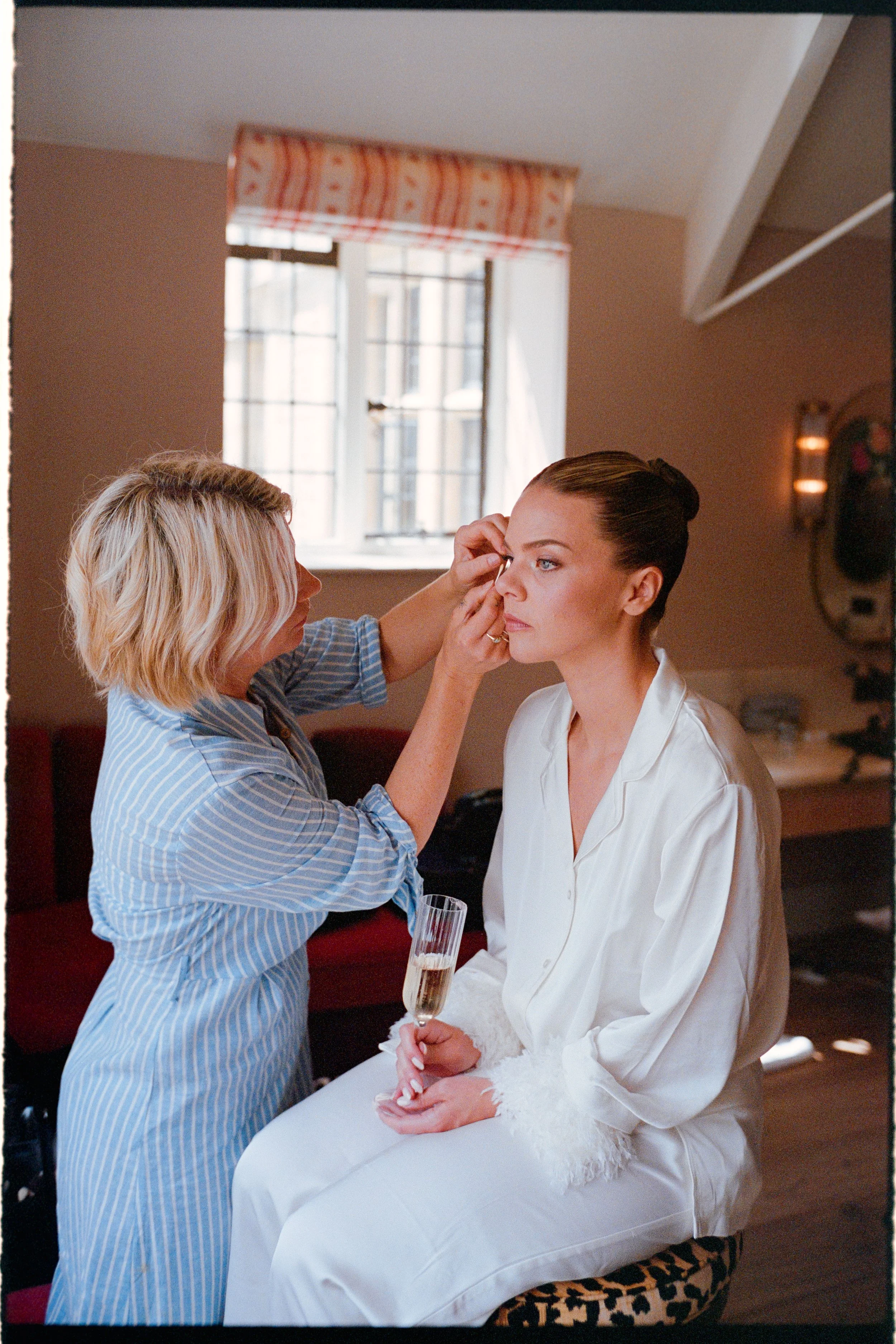 A woman having makeup applied while seated, with a glass of champagne in her hands, in a warmly lit room with a window in the background.