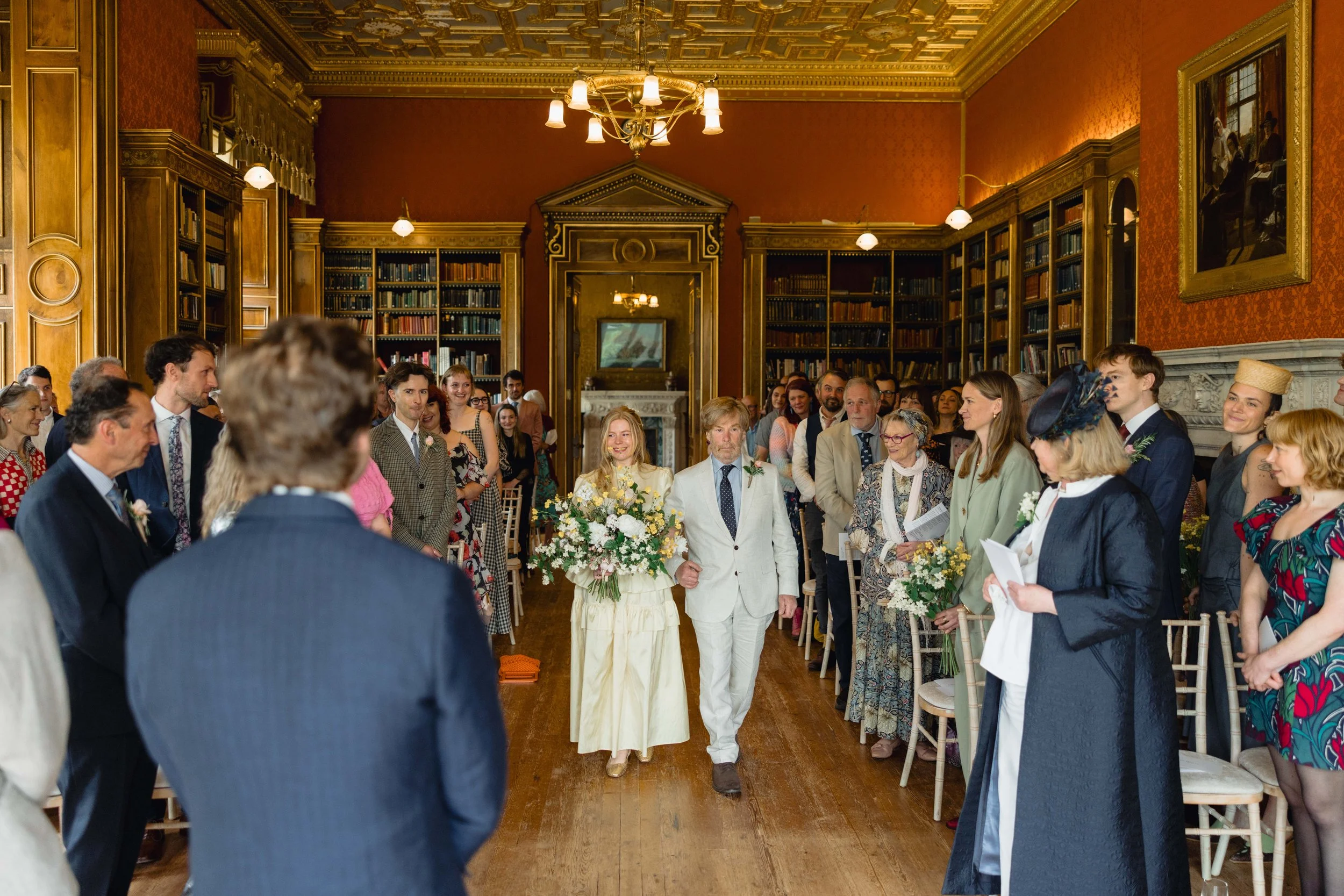 Wedding ceremony in an elegant, book-lined room with red walls and ornate woodwork. A bride in a cream dress is walking down the aisle accompanied by a man in a white suit. Guests are standing and seated on both sides, dressed in formal and vintage a