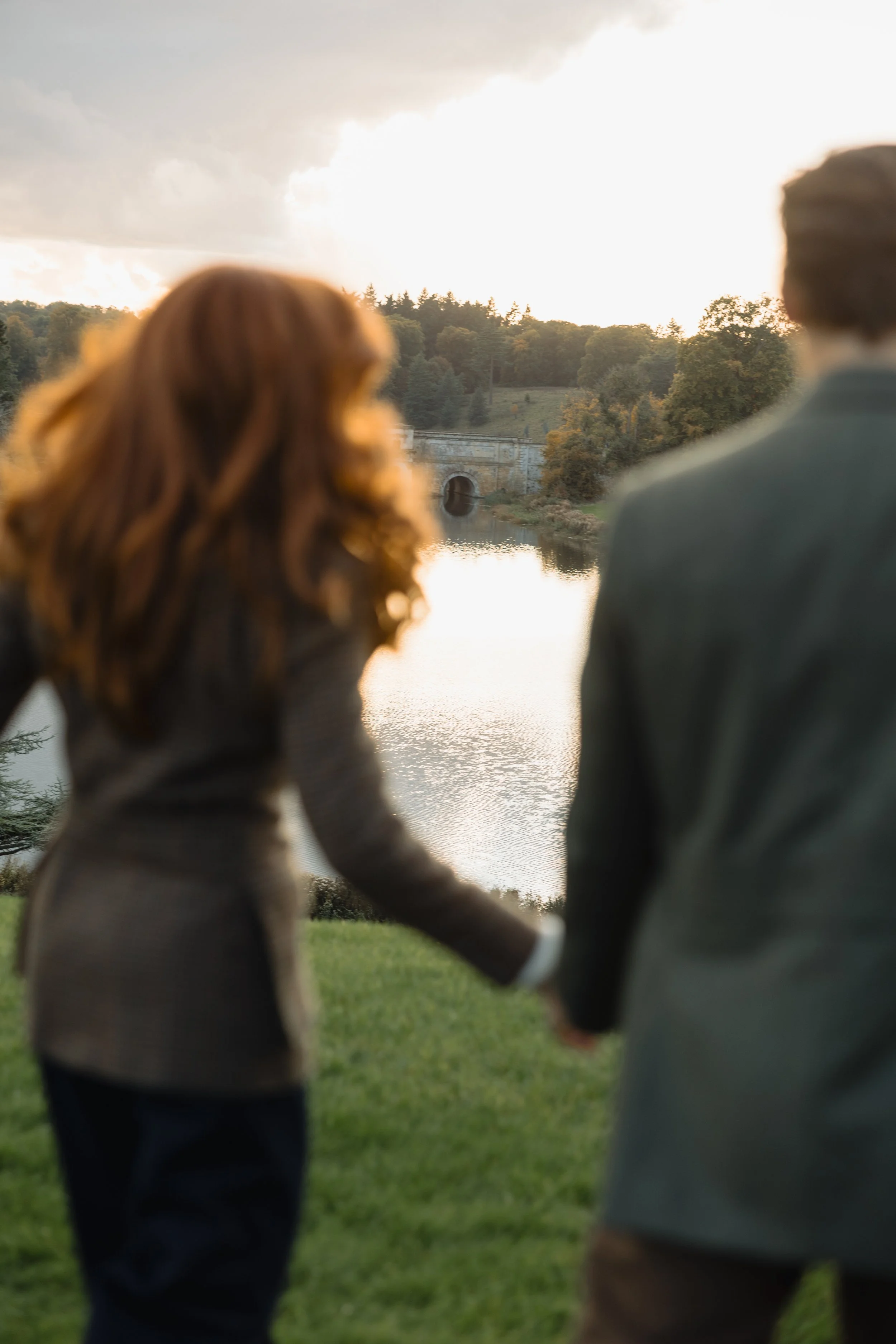 Two people are holding hands outdoors near a body of water, with trees and a bridge in the background, during sunset.