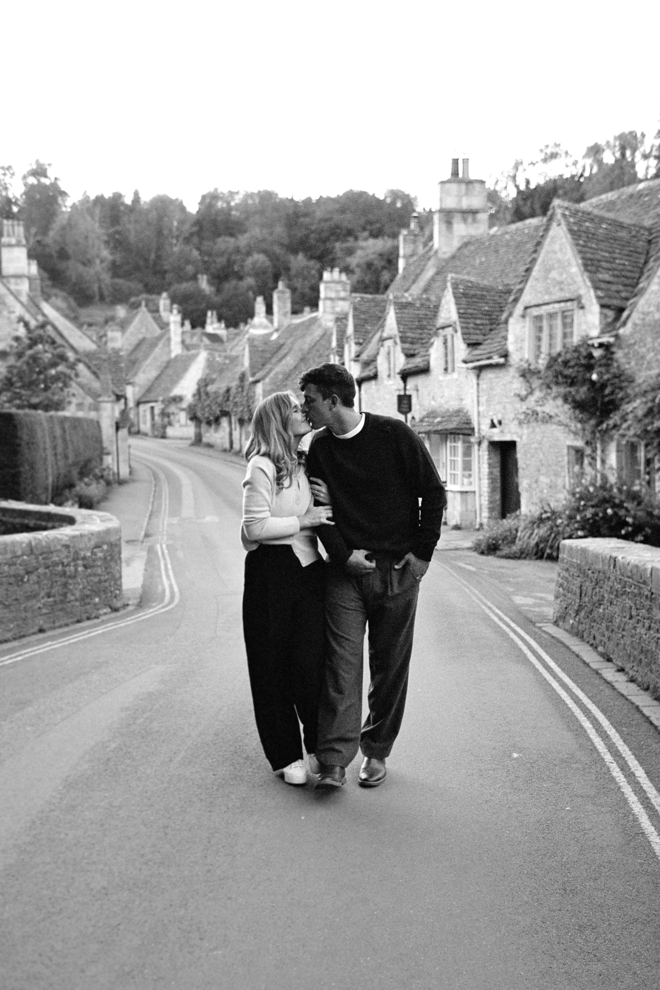 A young couple walking on a quiet, curved street in a small village, leaning in for a kiss. The street is lined with old houses with steep roofs and chimneys, and there's greenery in the background.