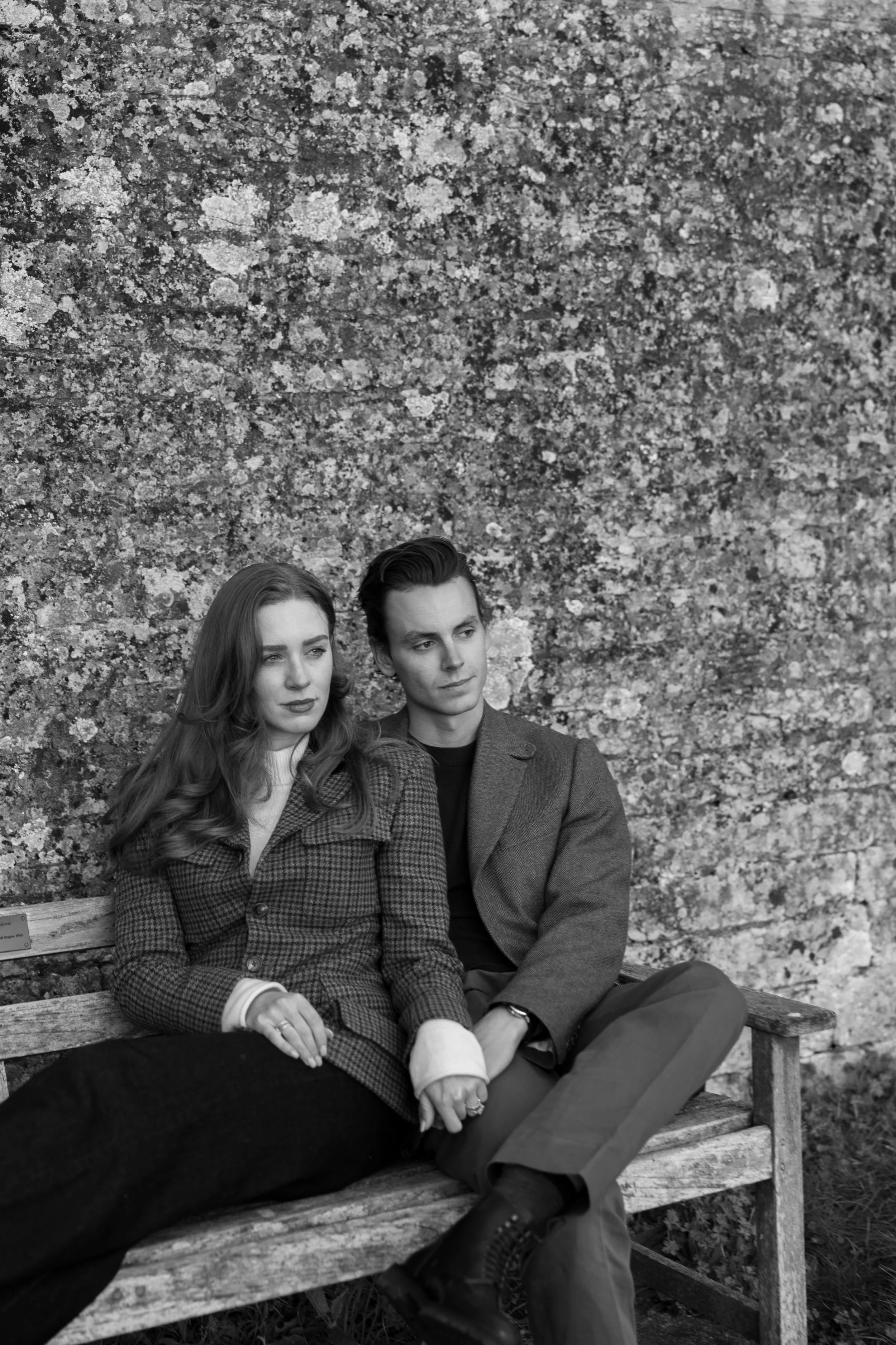 Black and white photo of a young man and woman sitting on a rustic wooden bench against a textured outdoor wall, holding hands and gazing pensively into the distance.