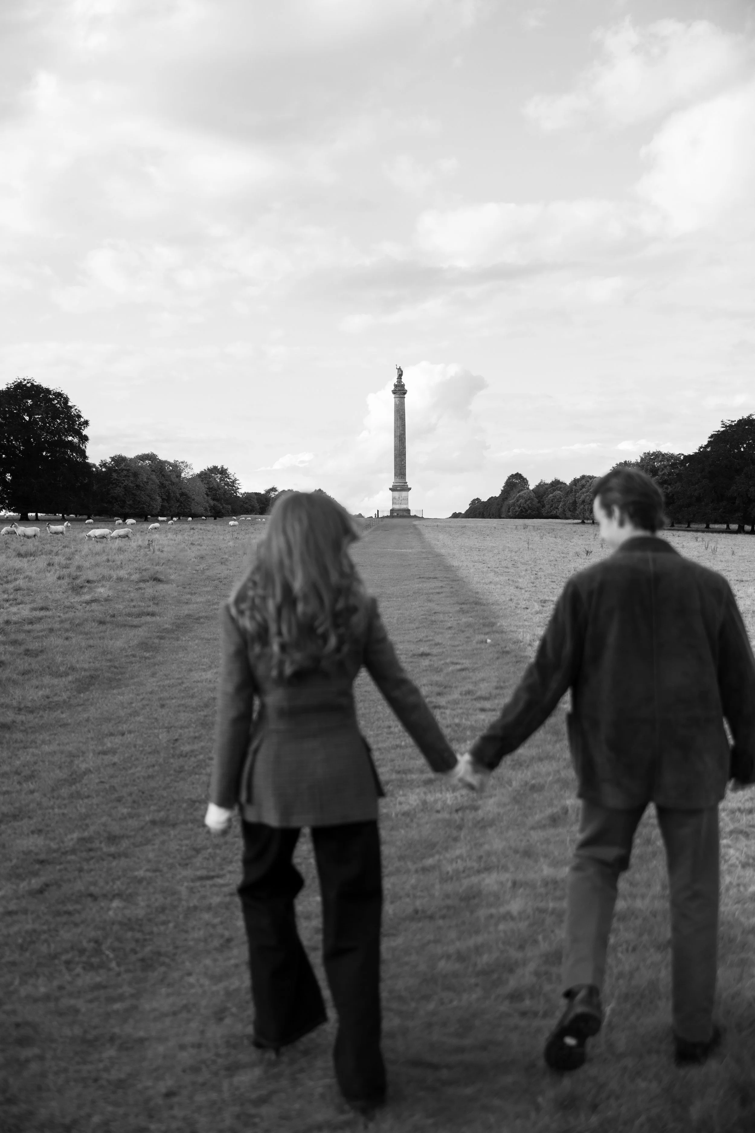 A black-and-white photograph of a couple holding hands, walking towards the Arc de Triomphe in the distance, with trees on either side and clouds in the sky.