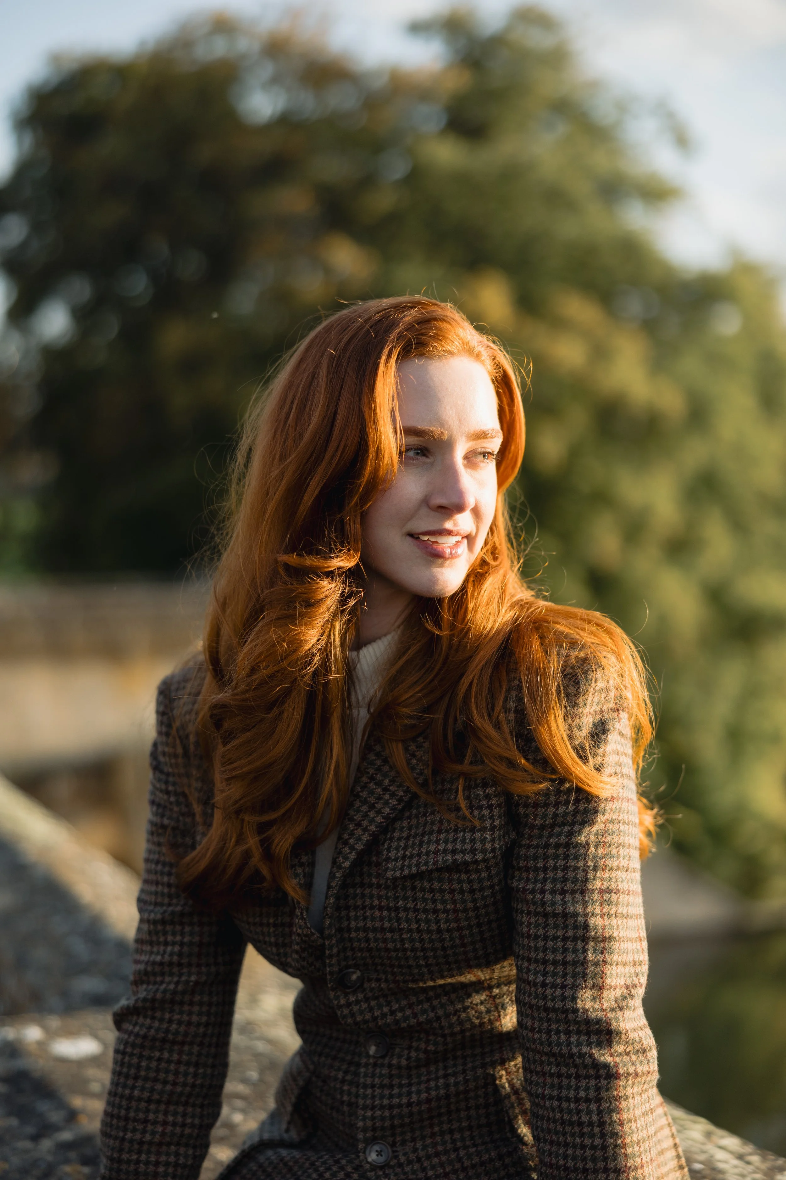 A young woman with long red hair styled in loose waves, wearing a checkered blazer, is outdoors near a body of water with trees in the background, during late afternoon sunlight.