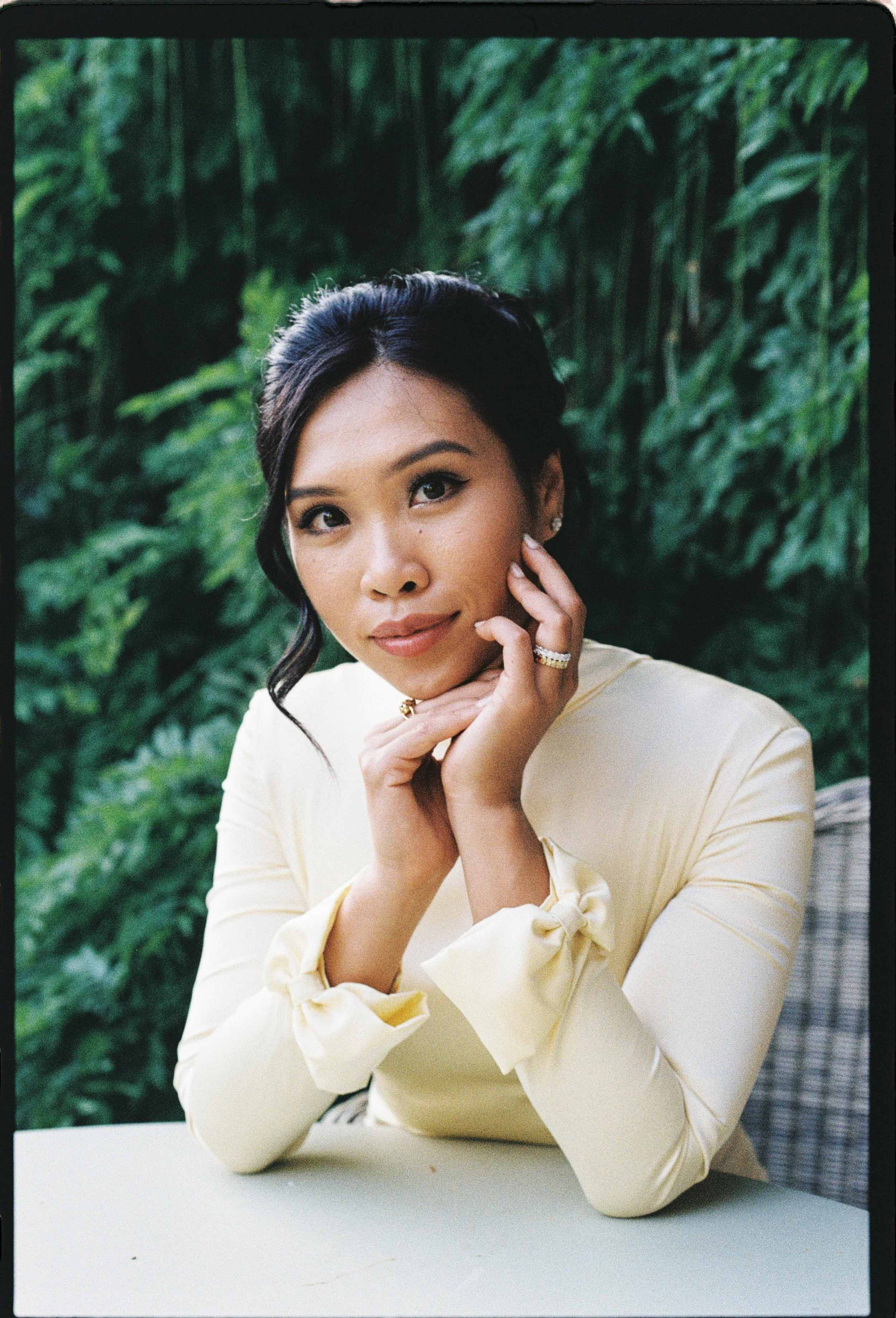 A woman with dark hair styled in waves, wearing makeup and pearl earrings, posing with her hand resting on her chin in front of lush green foliage.