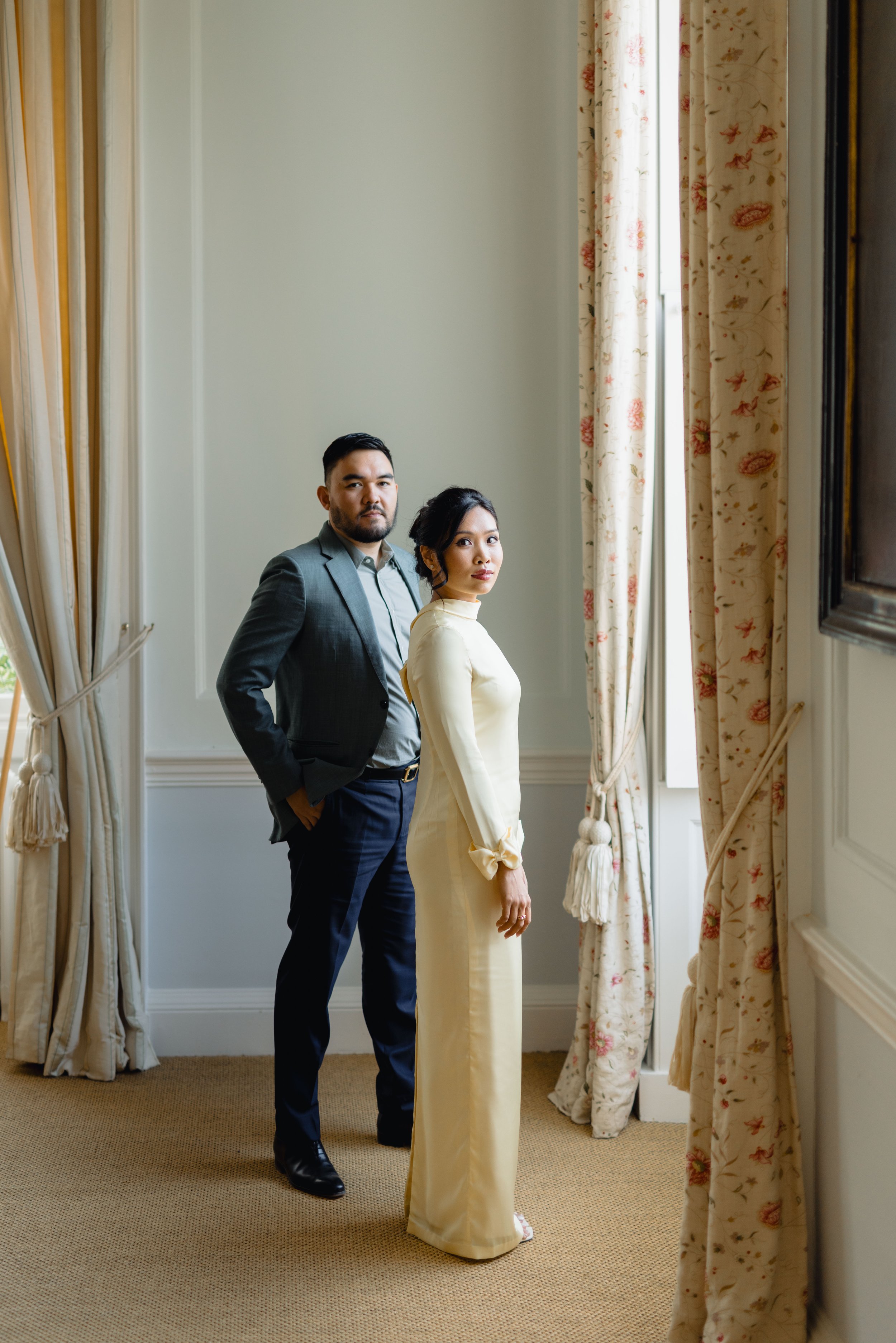 A man and woman standing inside a room with tall windows and floral curtains, looking at the camera.