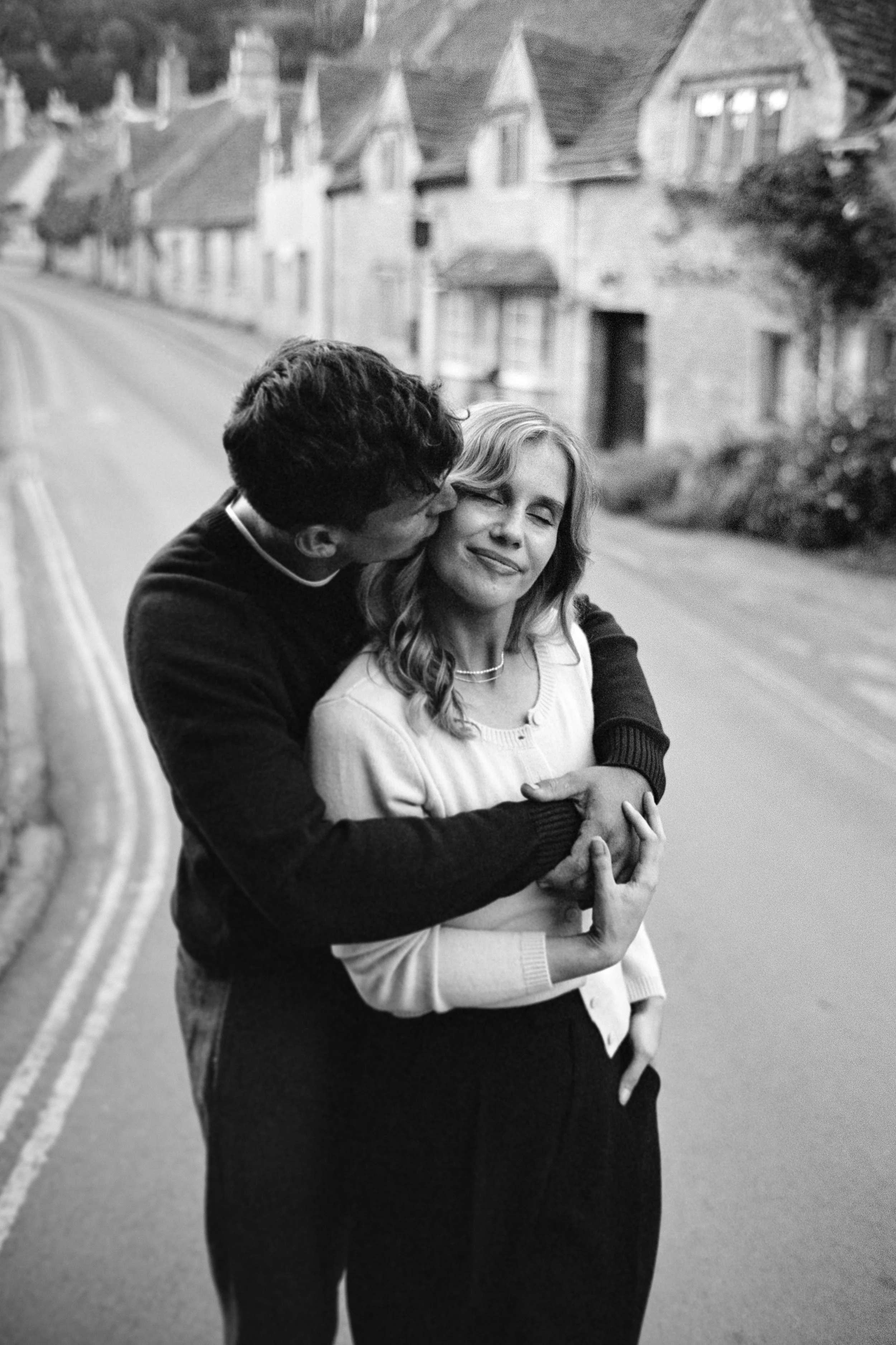 A black-and-white photo of a young man kissing a young woman on the cheek while holding her in a warm embrace on a quiet street with houses in the background.