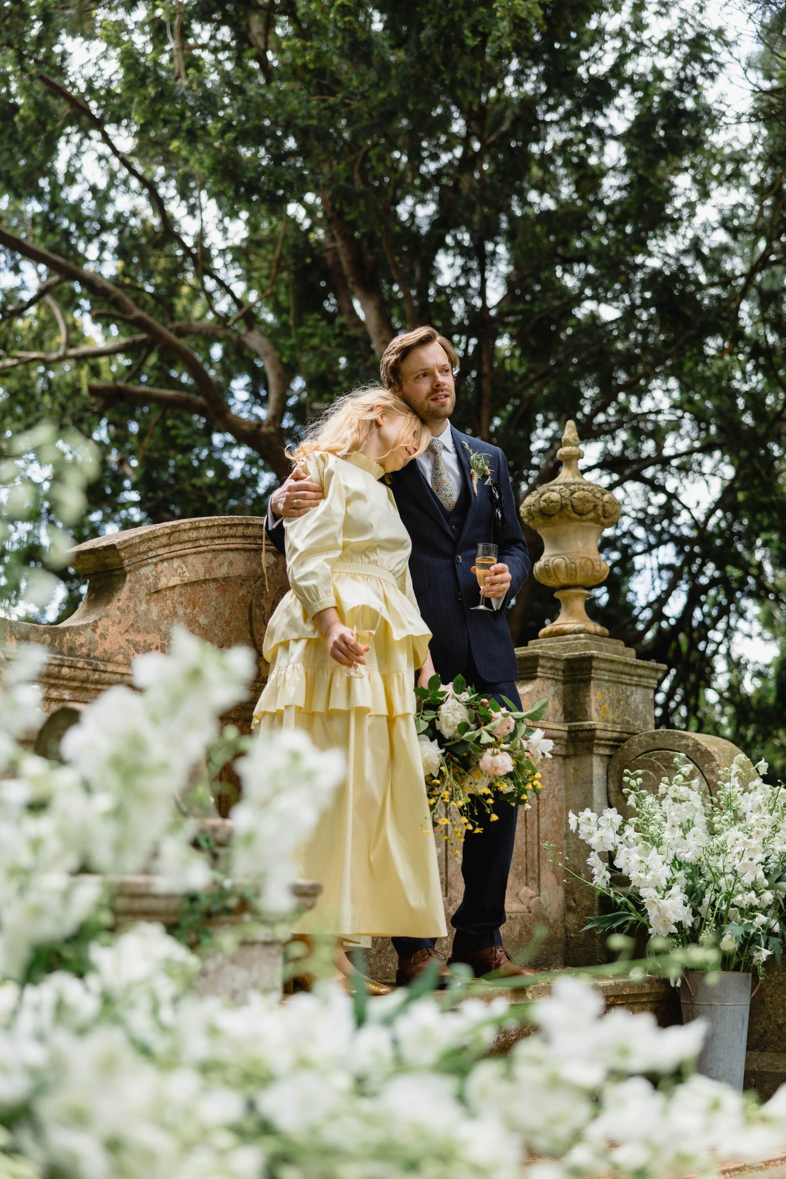 A man in a dark suit holding a glass of champagne hugs a woman in a yellow dress, who is resting her head on his shoulder, at an outdoor event surrounded by white flowers and greenery.