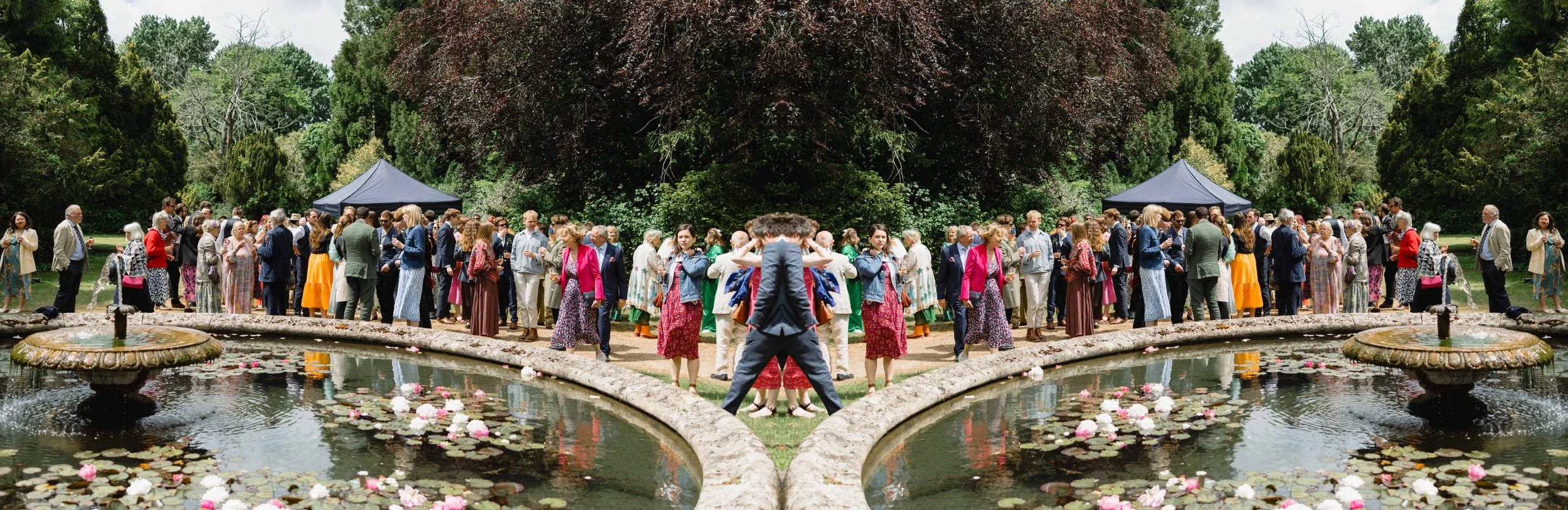 A large outdoor gathering of people in colorful clothing around a landscaped garden with ponds, fountains, and trees.