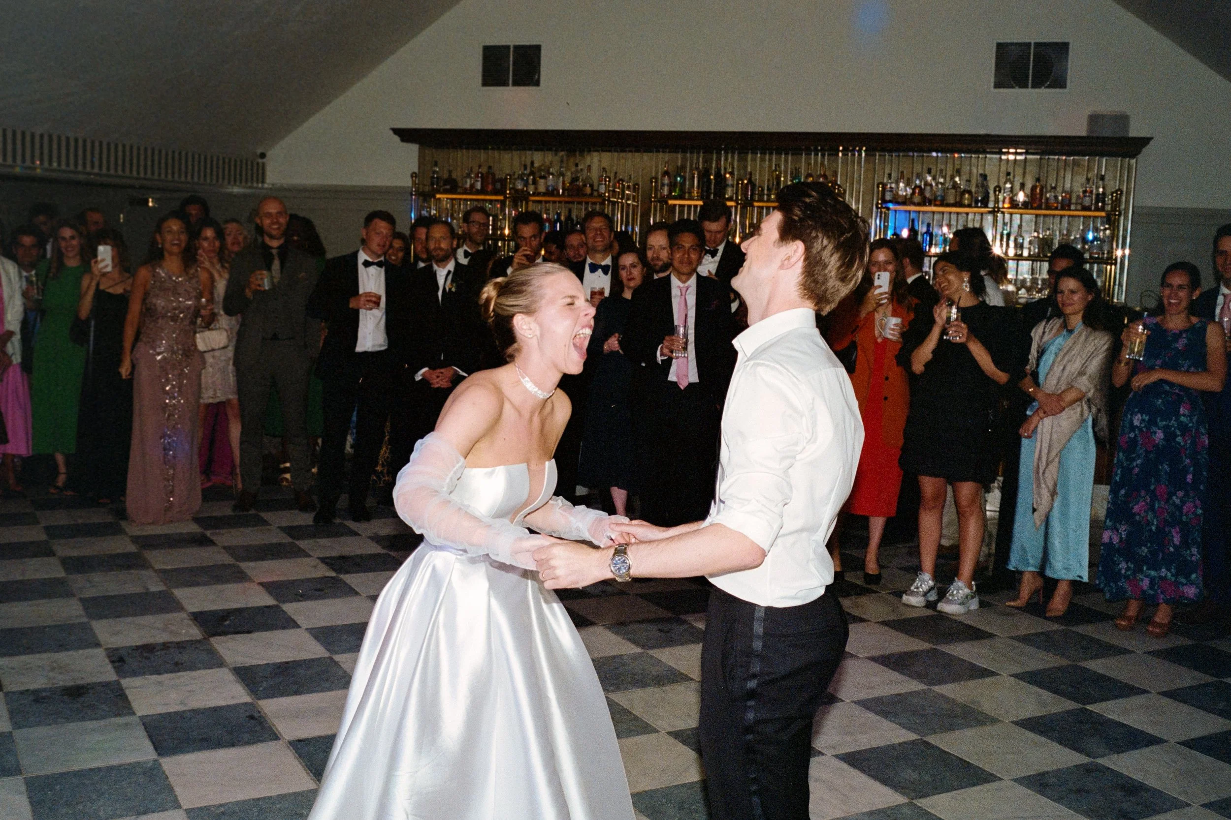 A bride and groom are dancing together at their wedding reception, surrounded by guests watching and taking photos.
