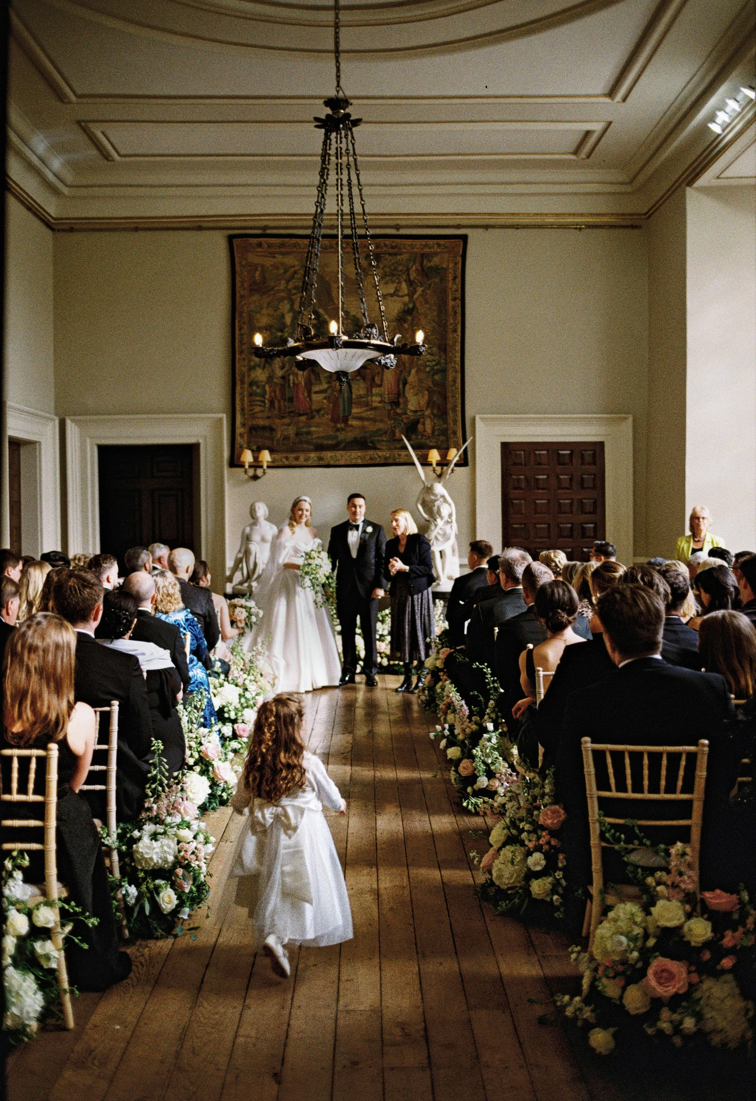 A wedding ceremony taking place in an elegant, historic room with high ceilings, chandeliers, and classical sculptures. The bride and groom stand at the front, surrounded by guests seated on either side, and a young flower girl runs down the aisle.