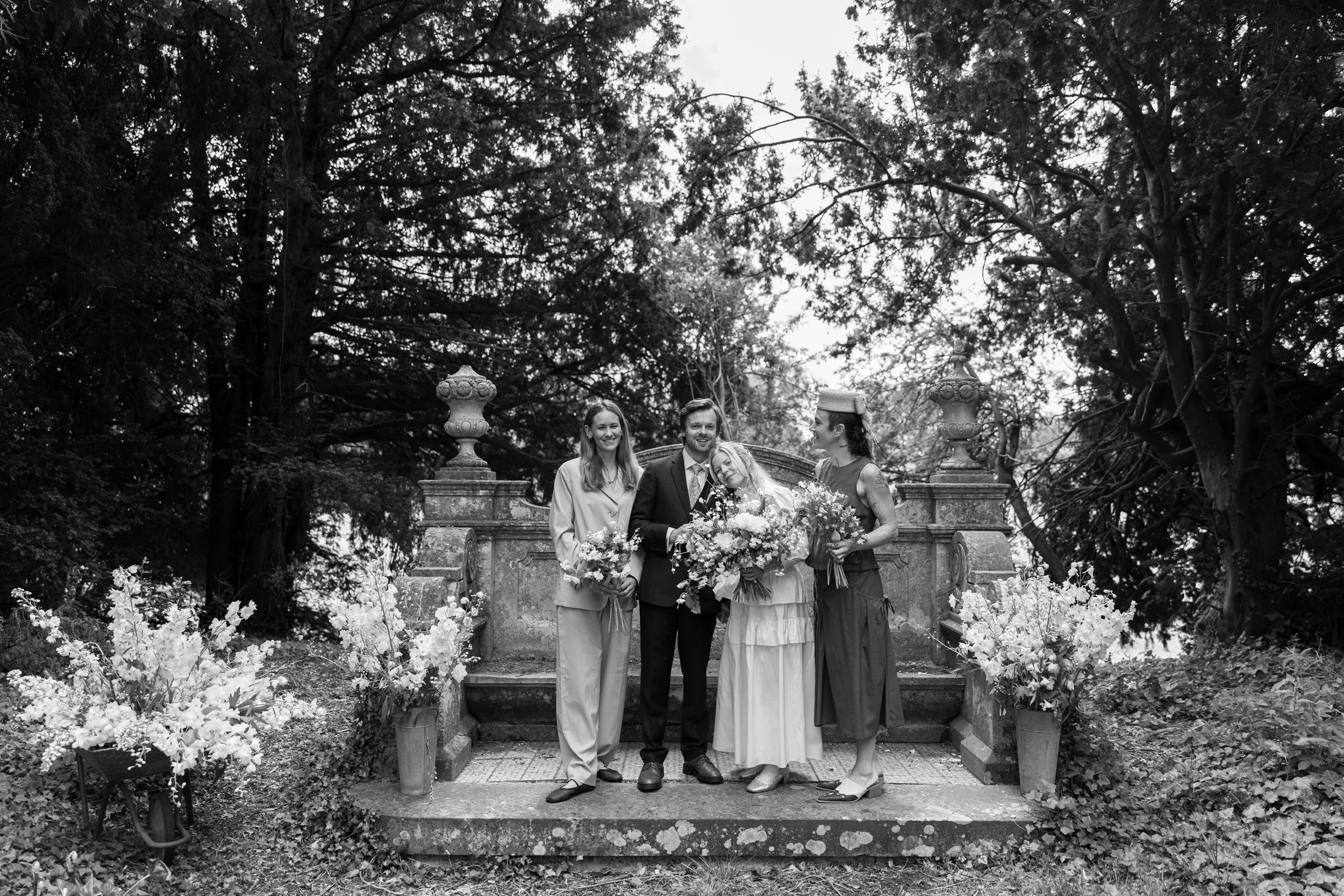 Black and white photo of four people at a wedding, standing on a stone platform with an ornate stone balustrade, surrounded by trees and potted flowers.