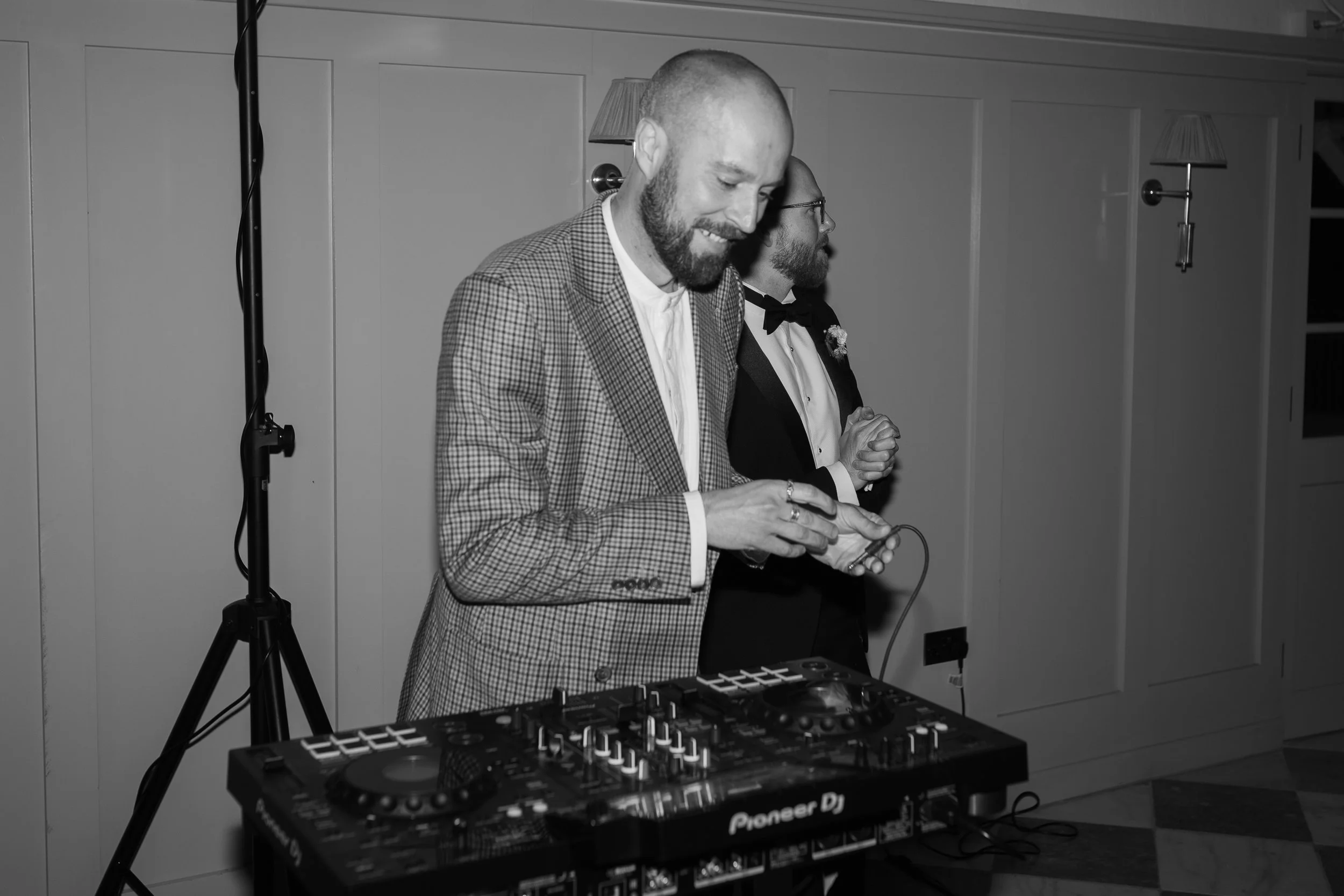 Two men in formal attire, one in a checked blazer and the other in a tuxedo, smiling and standing next to DJ equipment at an indoor event.